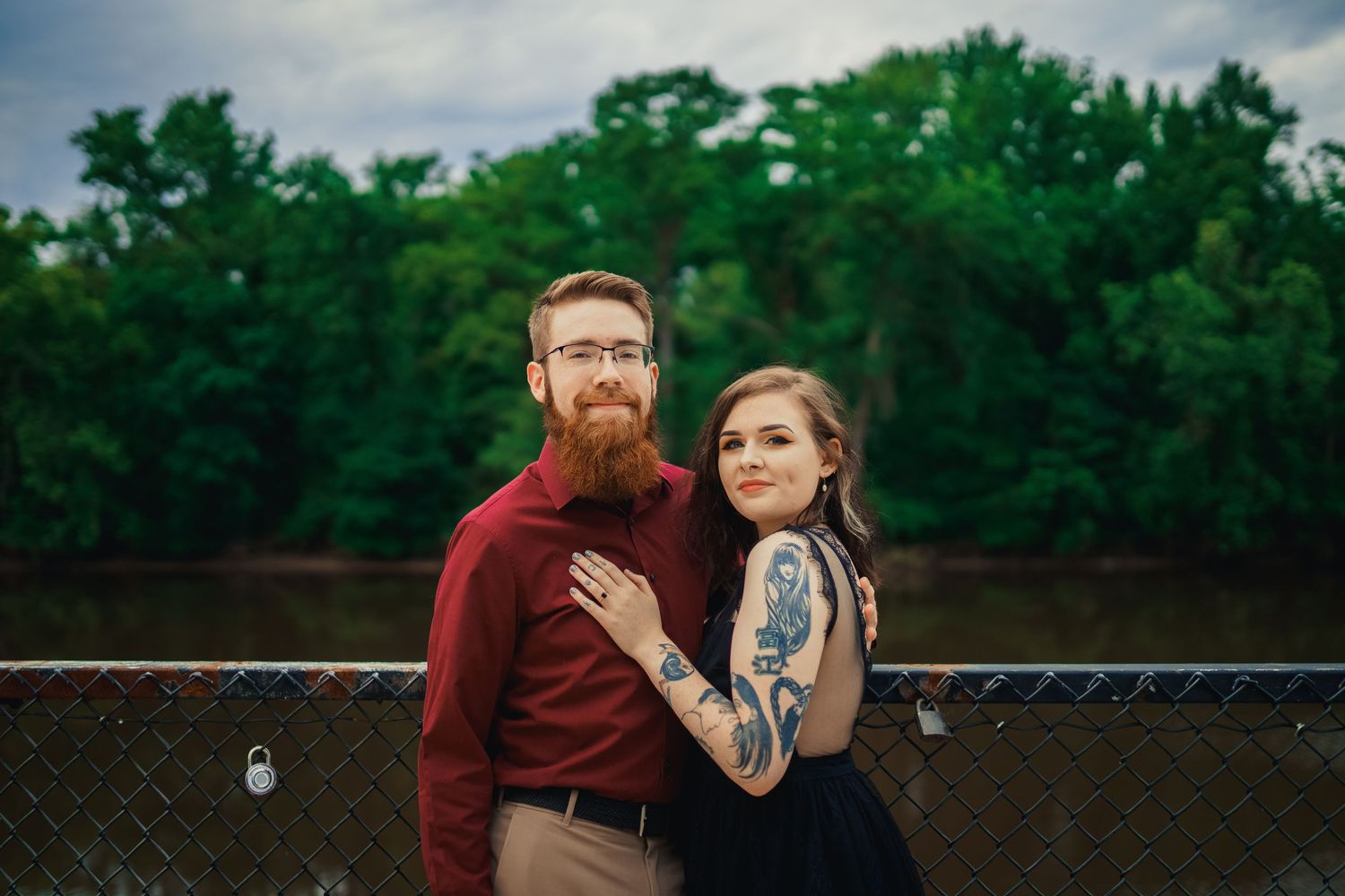 A couple in casual attire poses together against a fence showing off their colorful arm tattoos.
