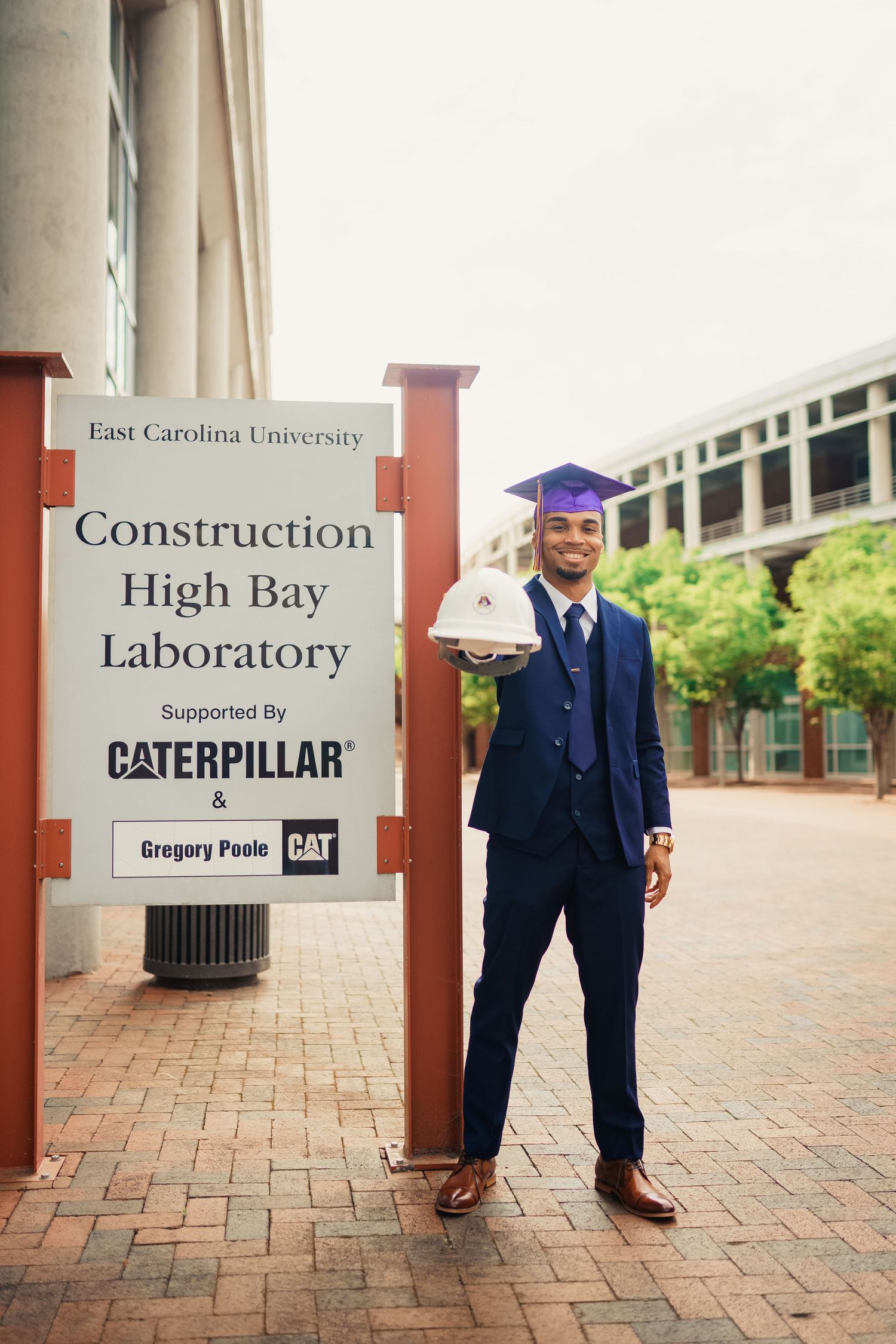Graduate in cap and suit stands next to East Carolina University Construction High Bay Laboratory sign supported by Caterpillar.