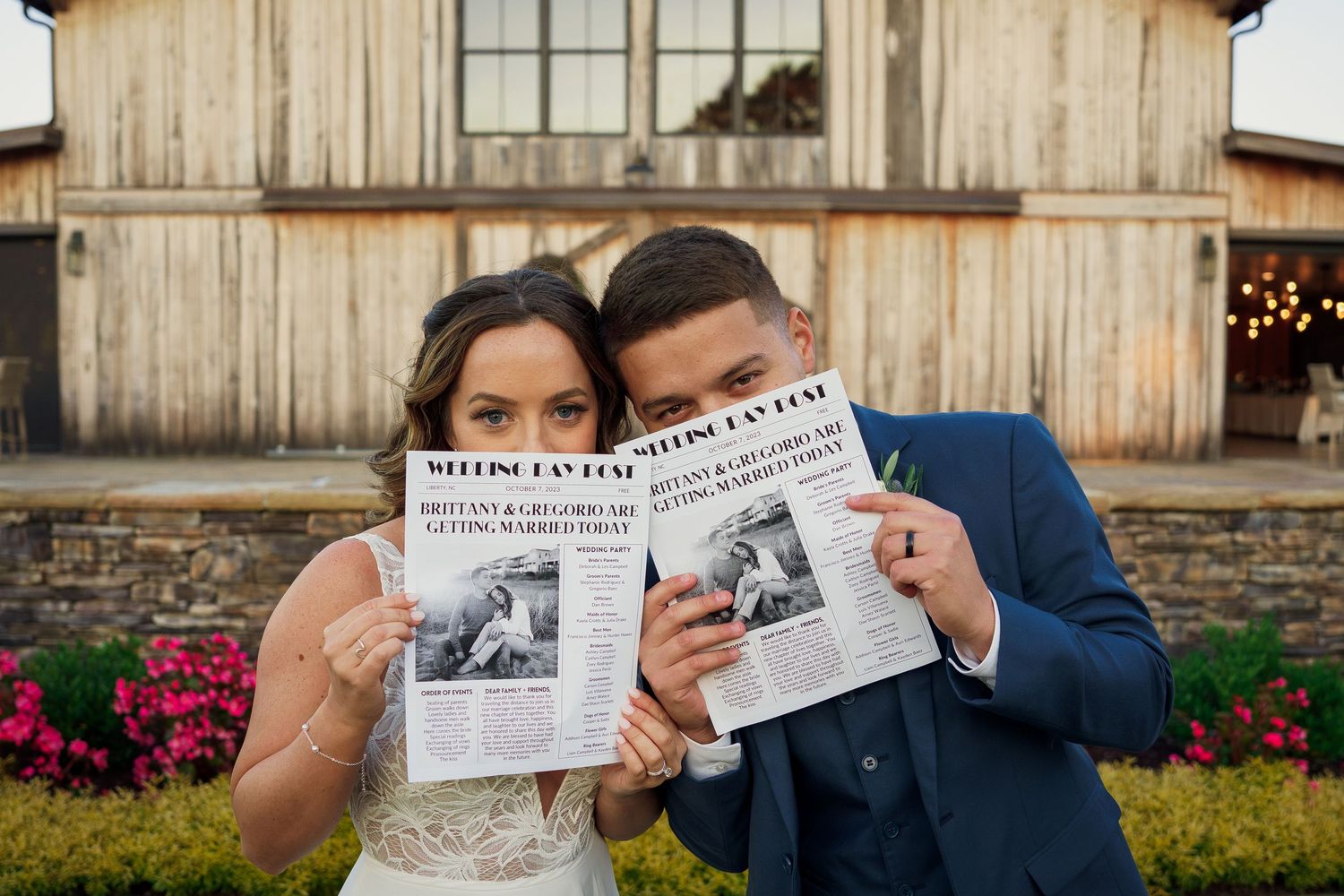 A wedding couple poses with newspapers in front of a rustic barn with flowering gardens.