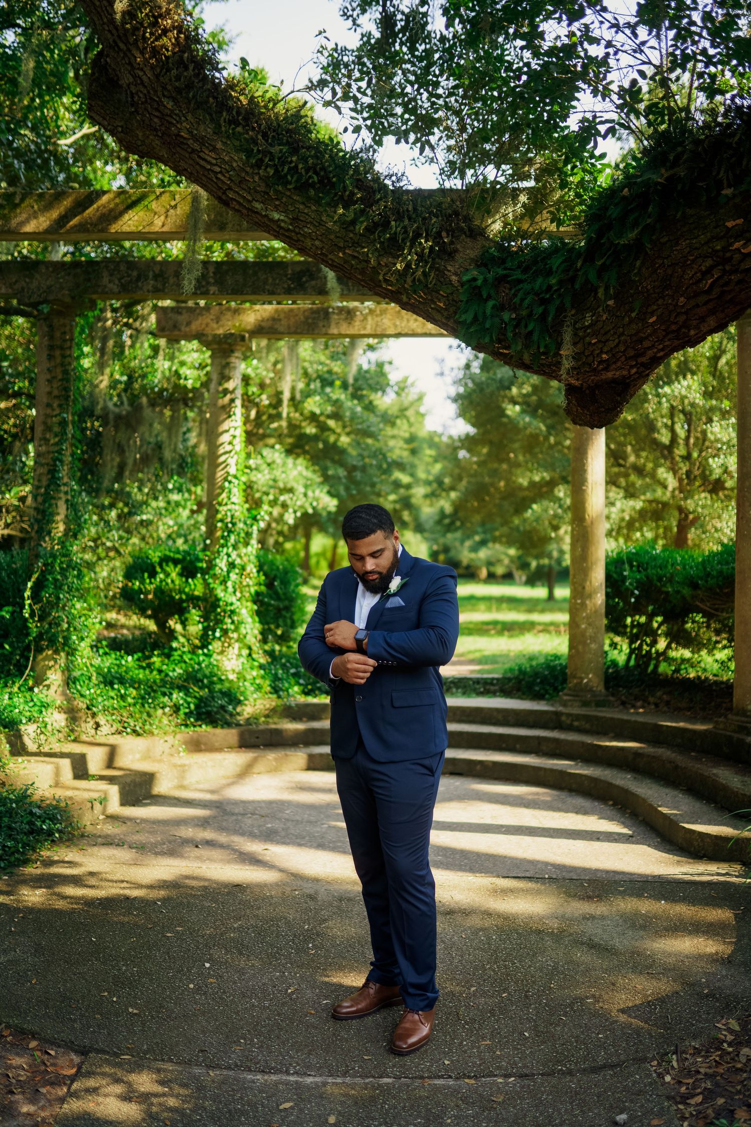 Formal portrait of groom adjusting suit jacket under garden pergola.