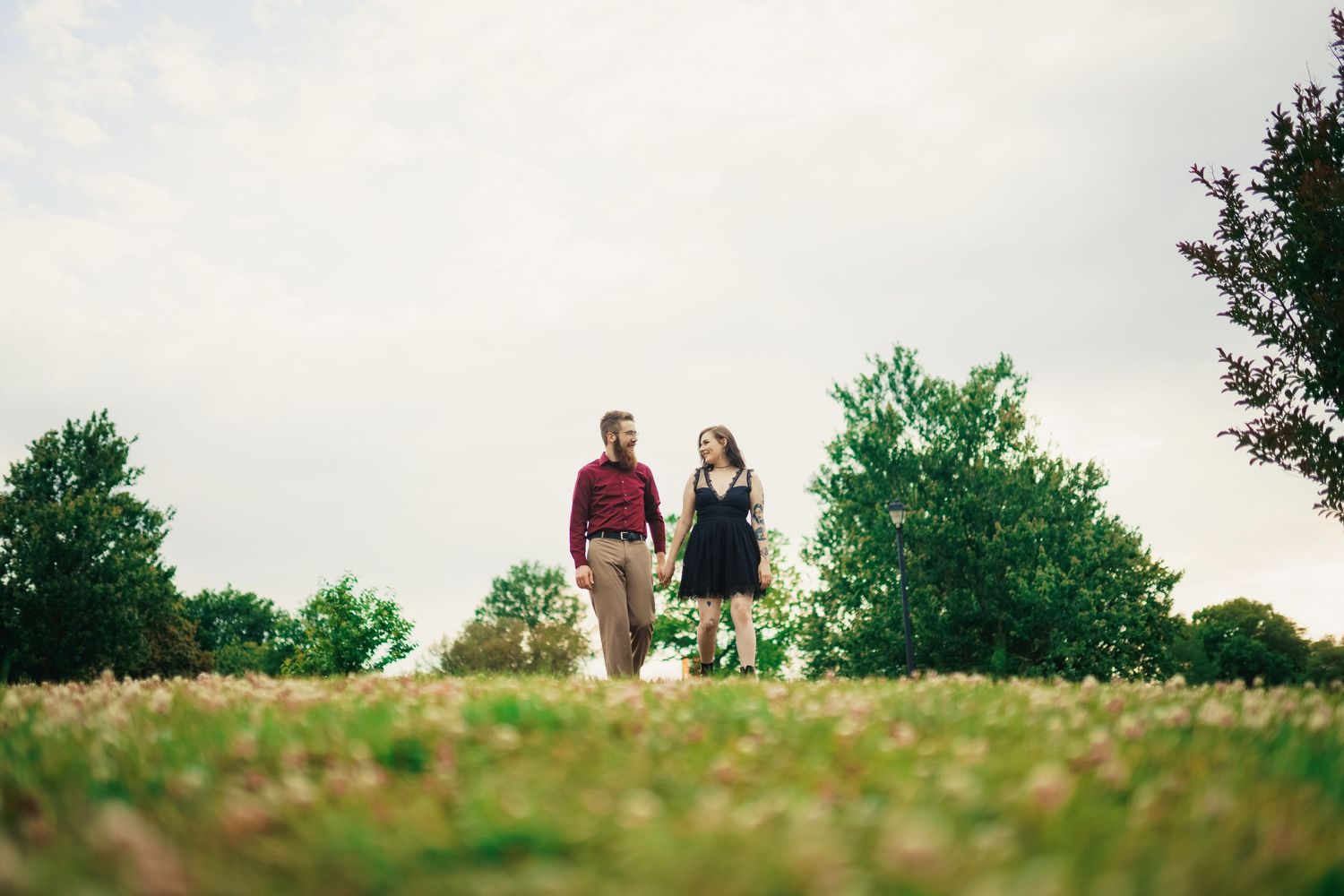 A couple takes a romantic stroll through a flowering meadow beneath cloudy skies.