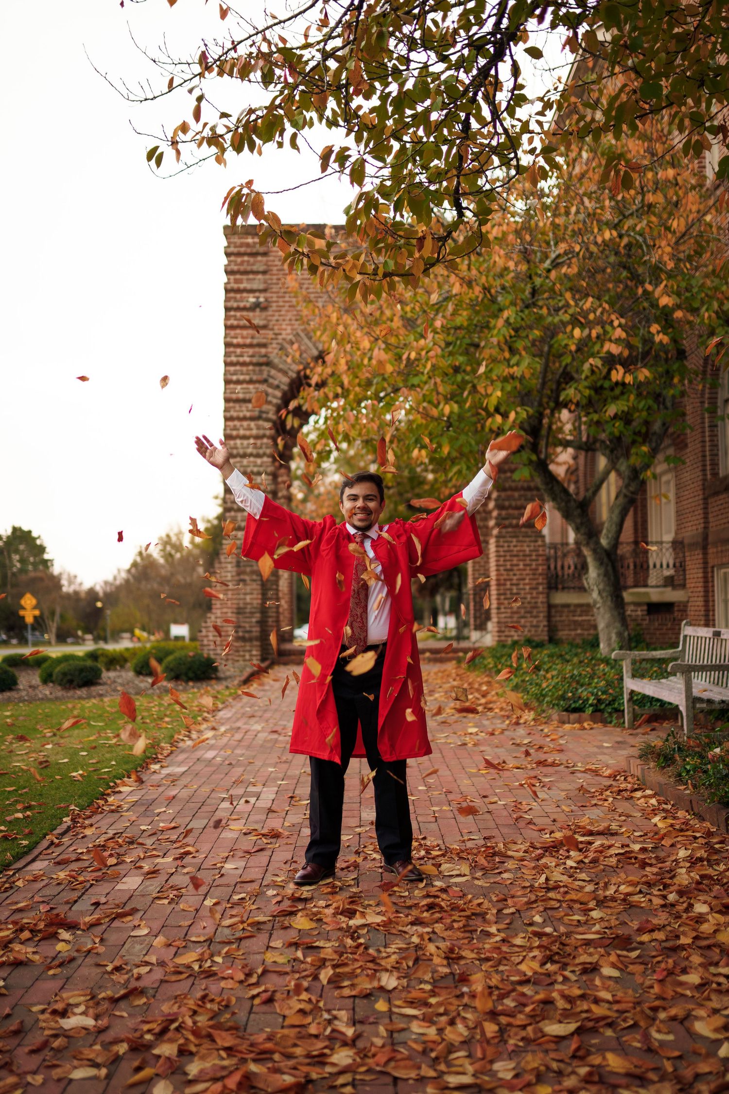 Graduate celebrates on brick pathway with falling autumn leaves wearing red graduation gown.