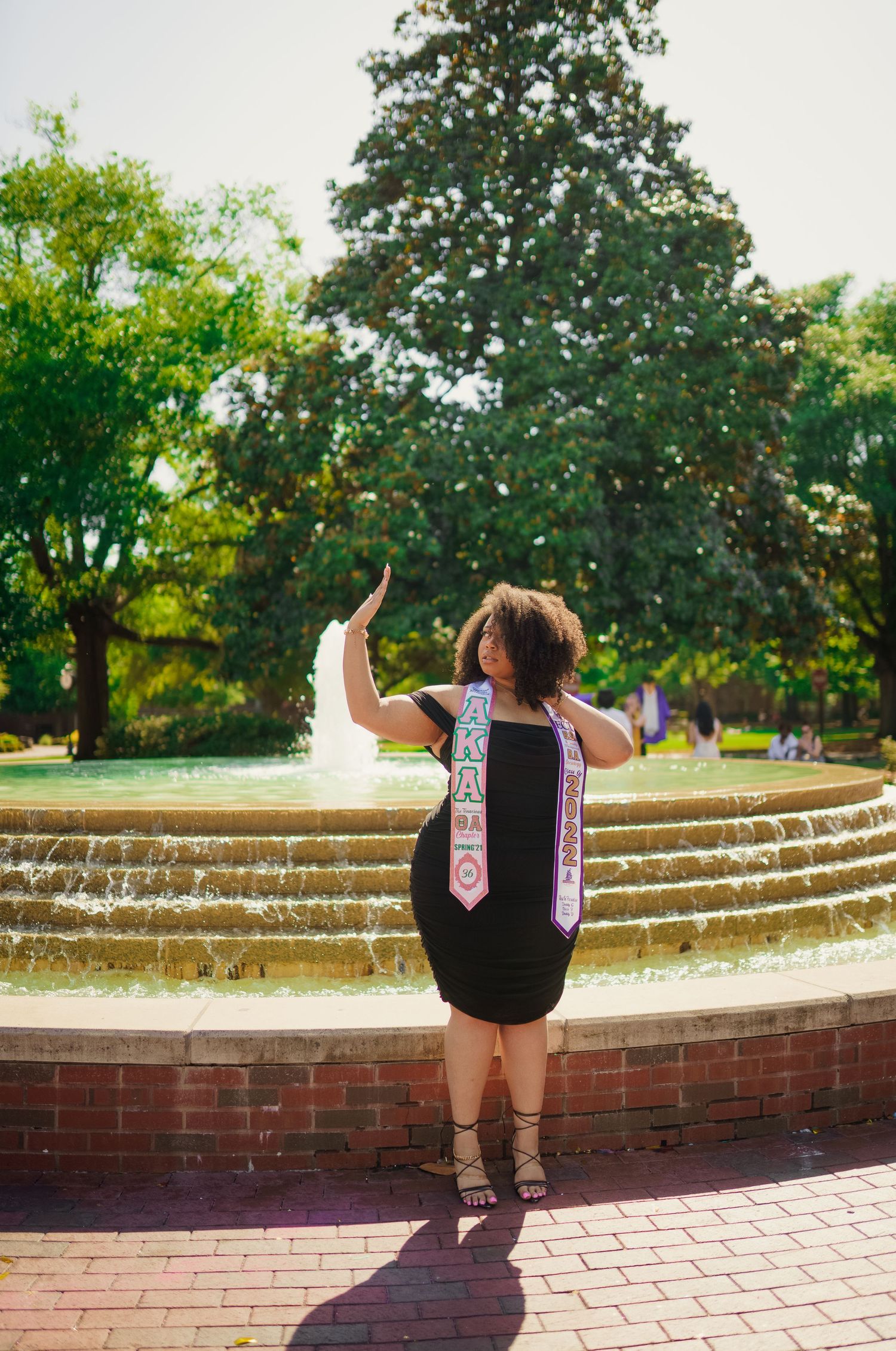 Graduate wearing honors cords celebrates by wright east carolina university fountain on brick path in sunny park setting with lush trees.