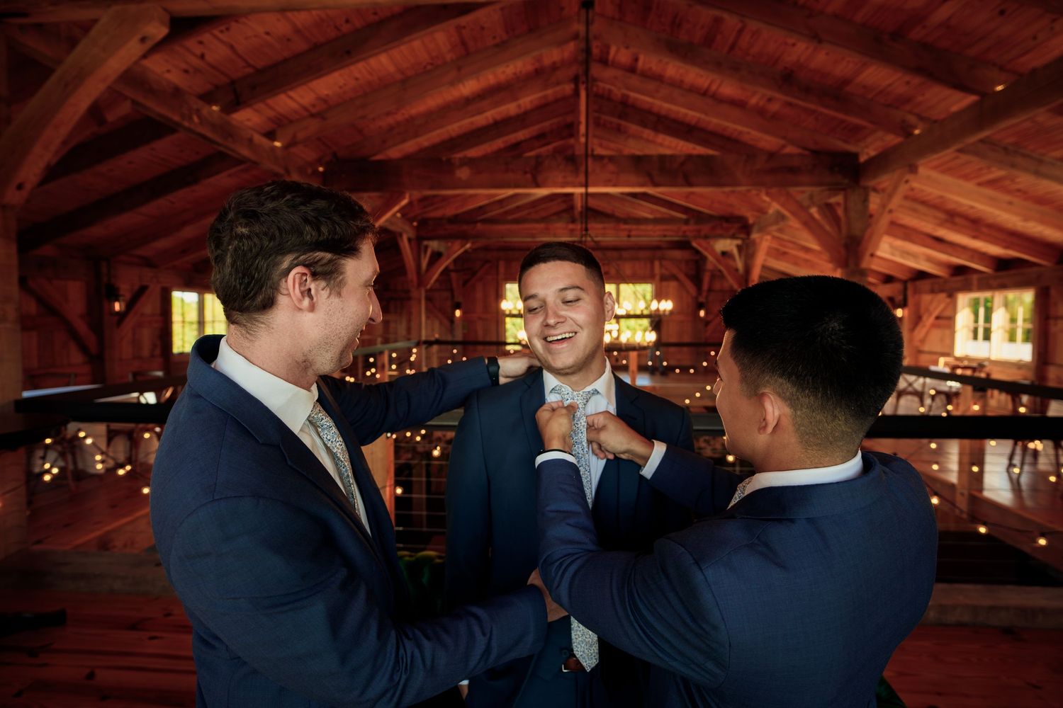 Three groomsmen in navy suits share a lighthearted moment in a rustic wooden barn venue.