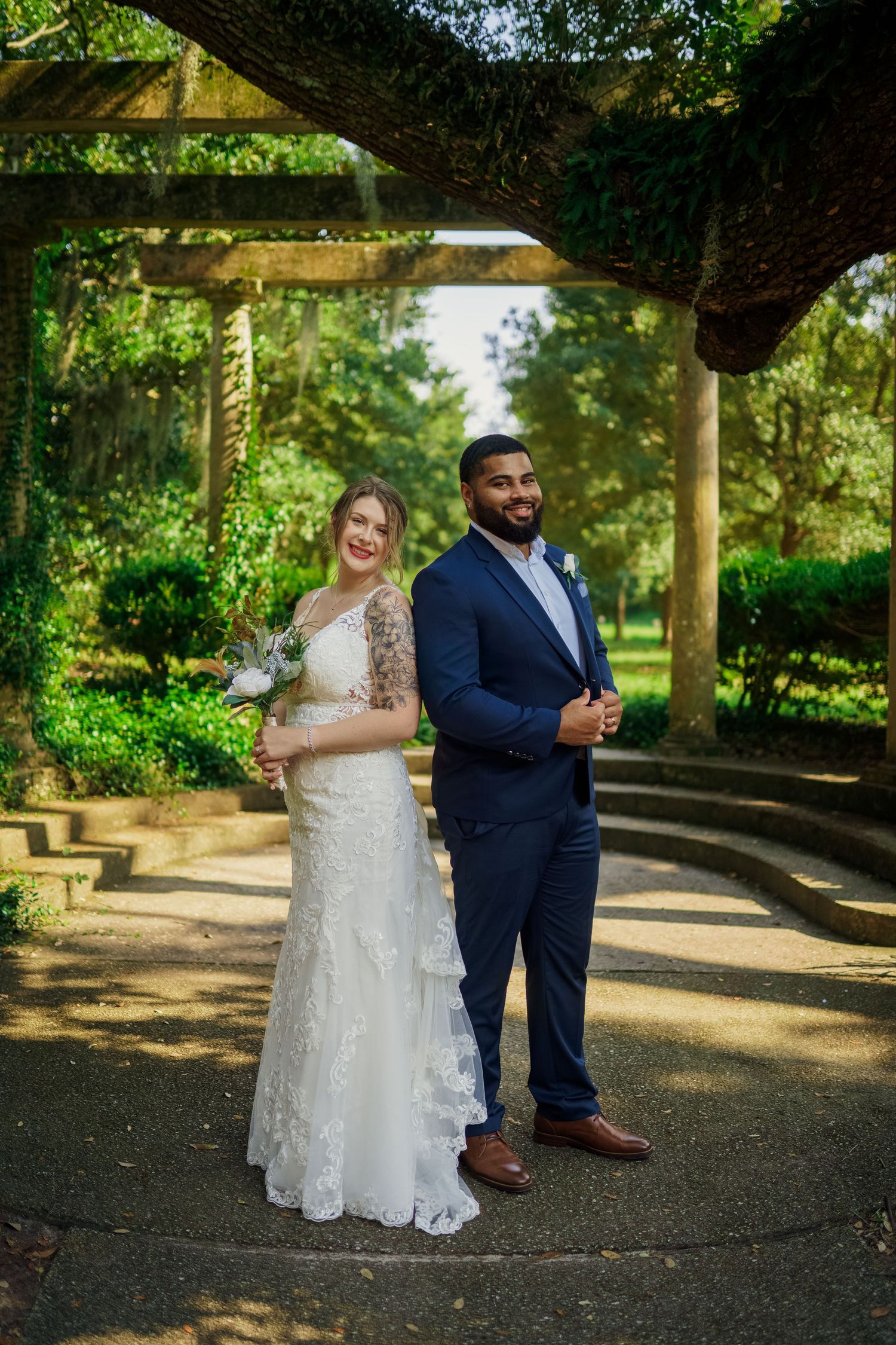 Couple in wedding attire pose back-to-back under garden archway on curved stone path.