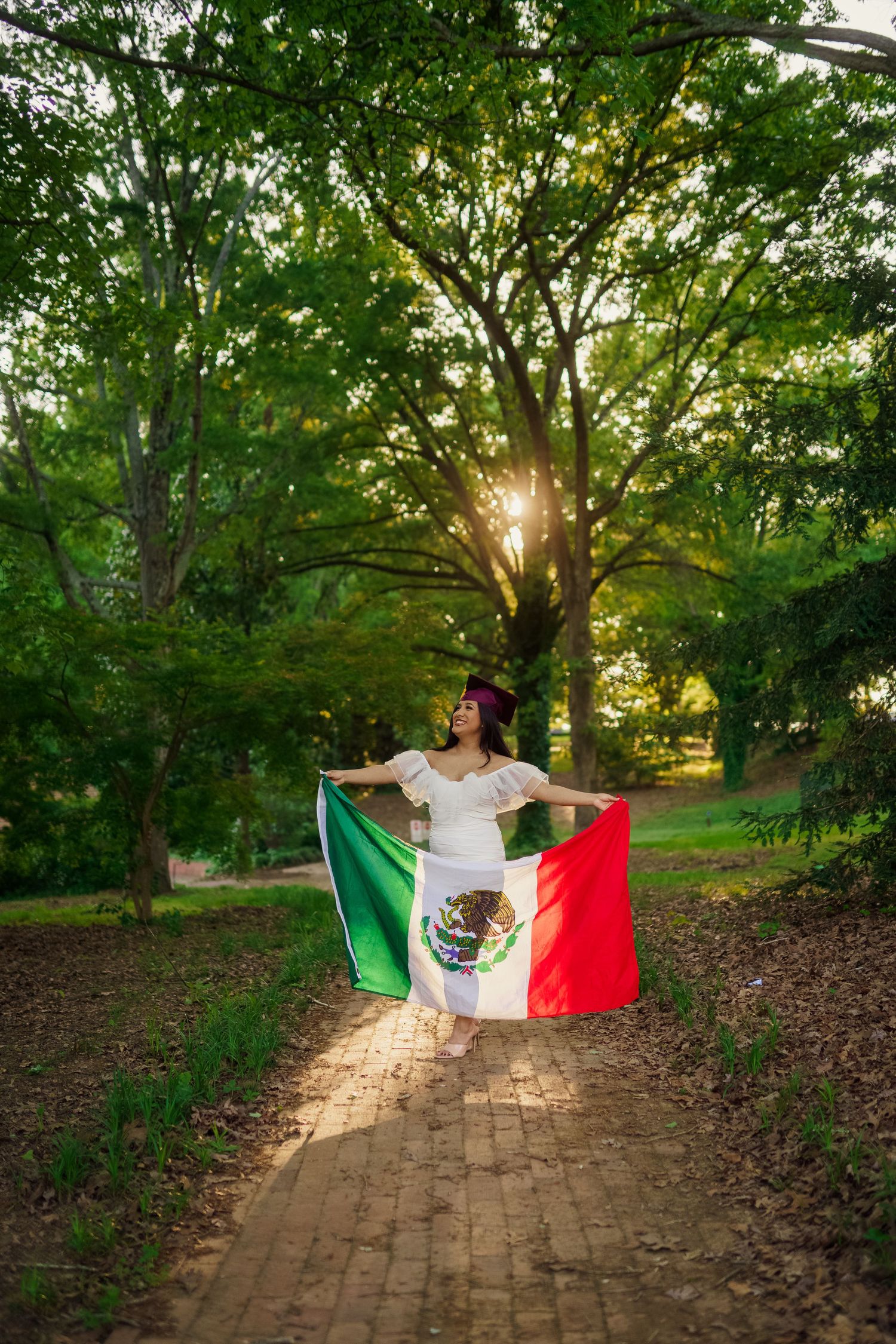 Surrounded by trees at sunset, a graduate poses with Mexican flag on brick pathway.