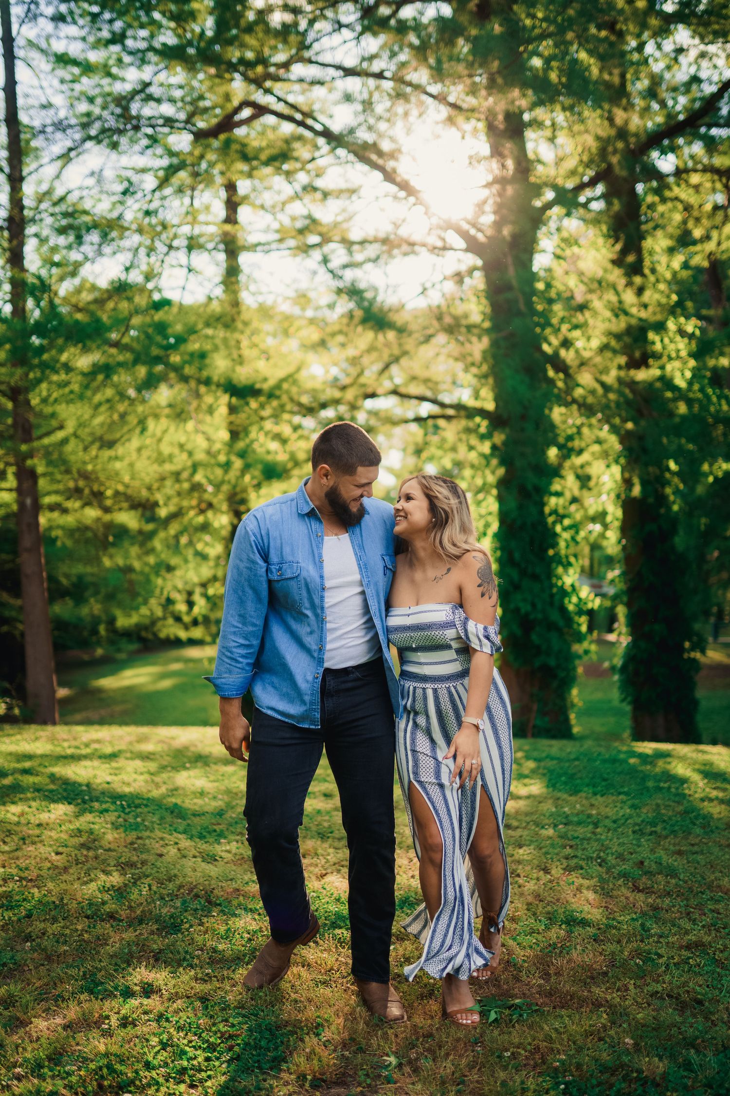 A couple shares an intimate moment while walking through a sun-dappled forest pathway.