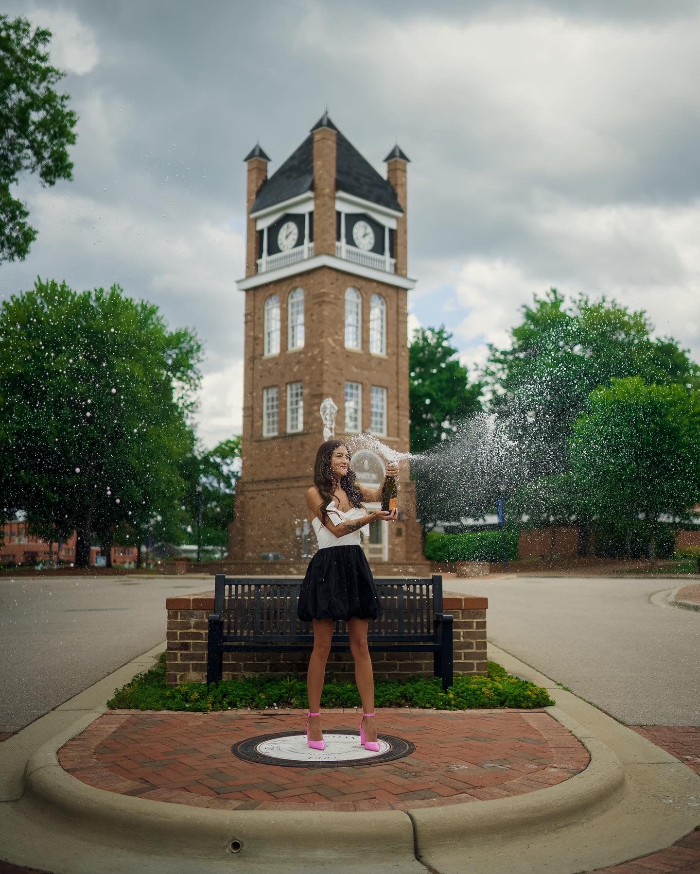 Clock tower stands tall against cloudy sky as Barton university graduate in black skirt celebrates on brick plaza. with champagne spray