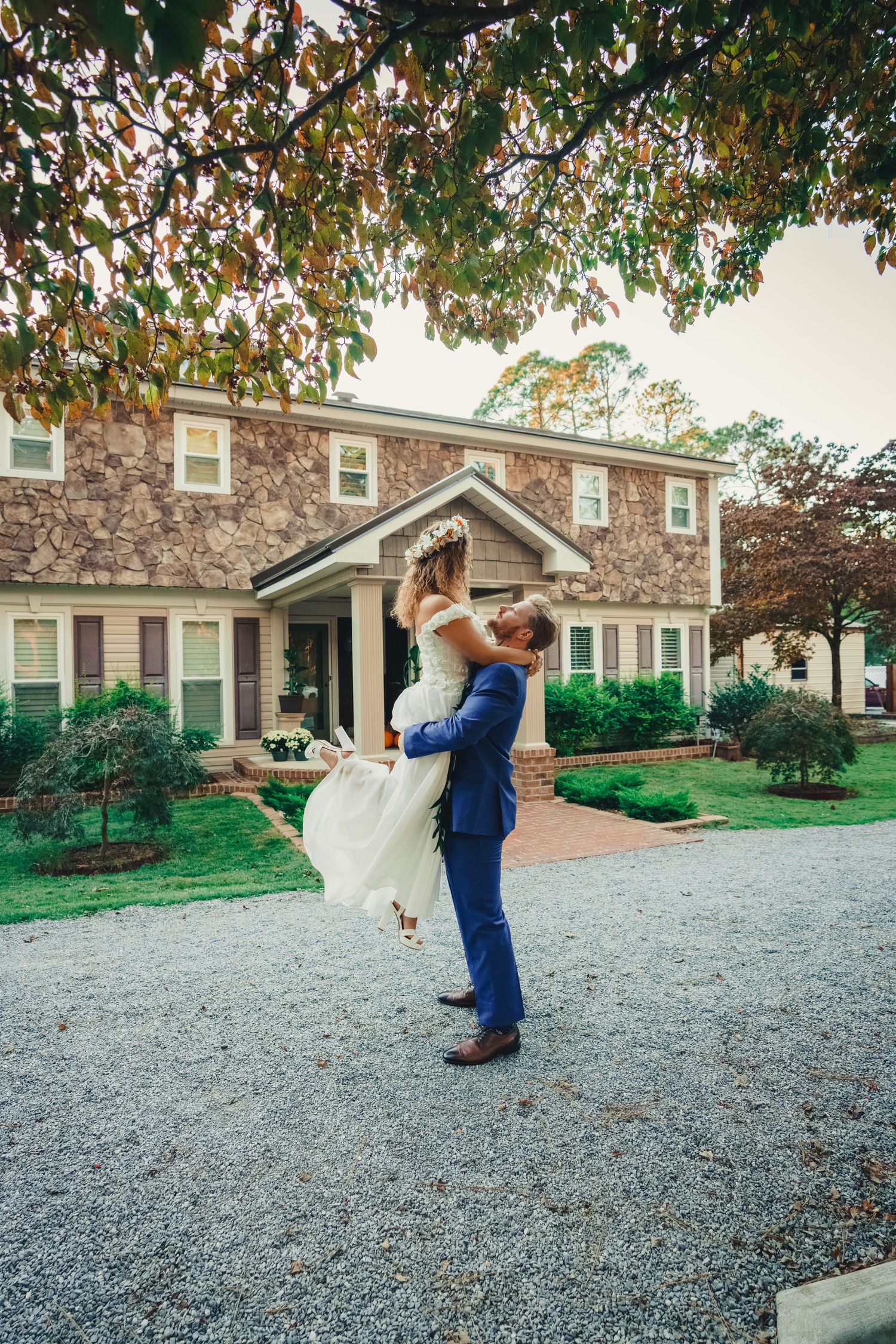 A romantic wedding moment captured outside a stone house as a couple shares a joyful lift on a gravel driveway. Joan Avilez Wedding photo