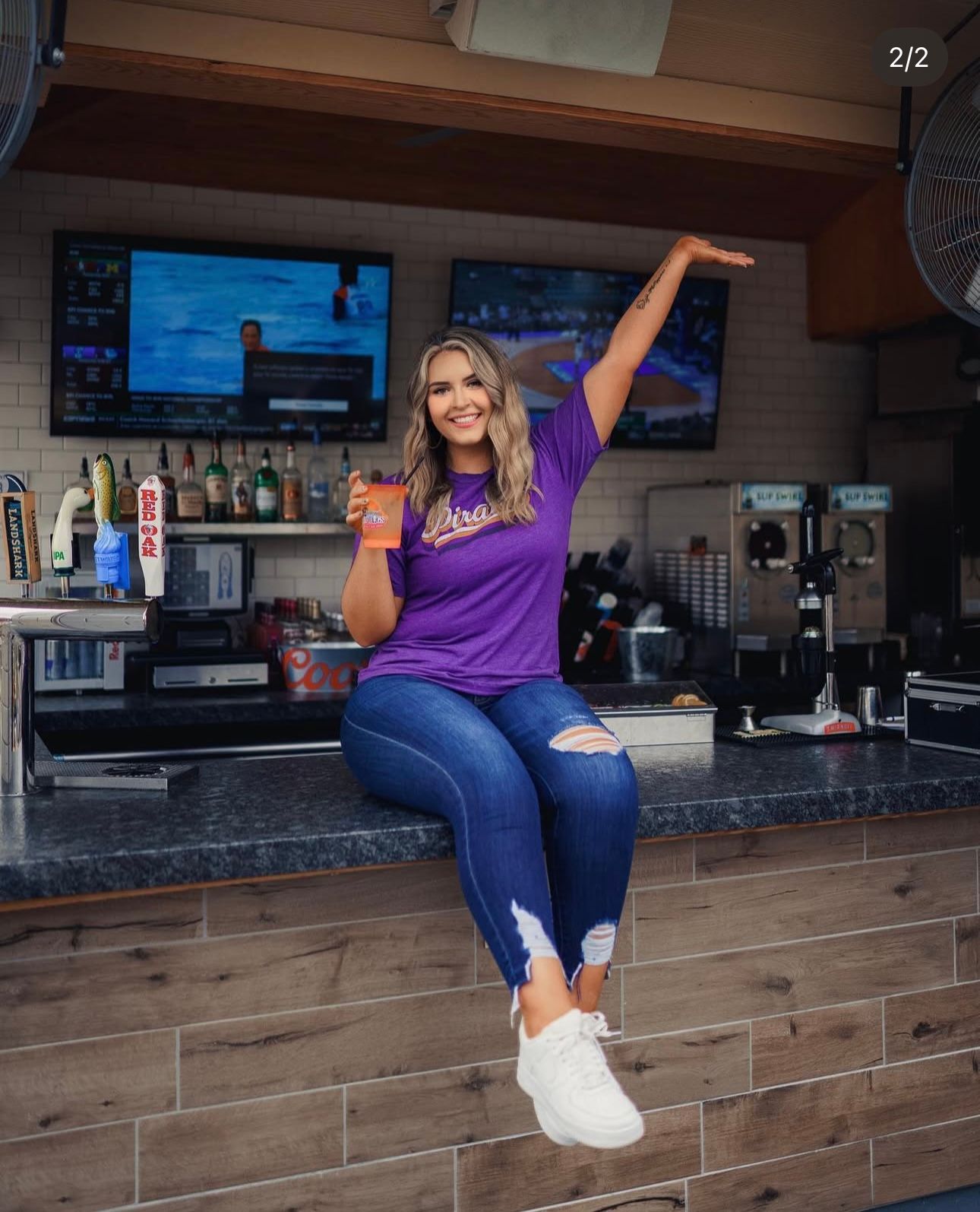 A person in purple sits casually on a bar counter at sup dogs with one arm raised in a celebratory pose.