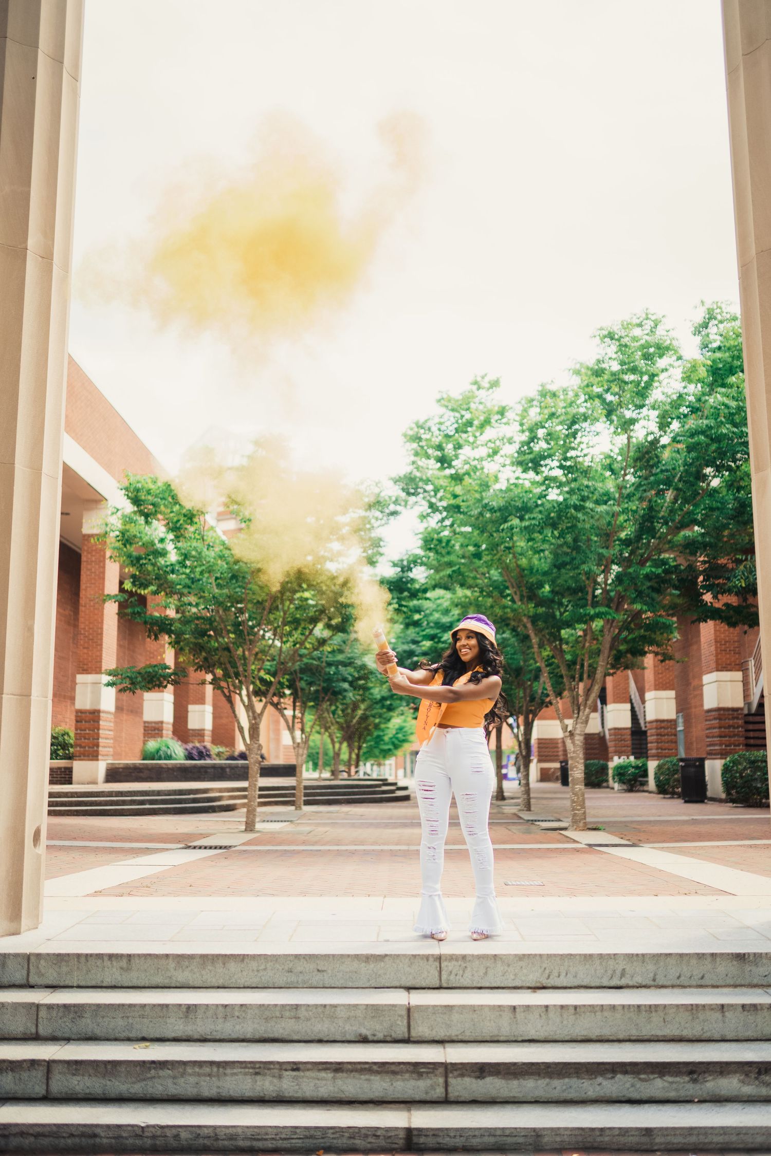 A ECU Graduate in white pants and yellow top stands on concrete steps in front of a brick building with green trees on a sunny day.