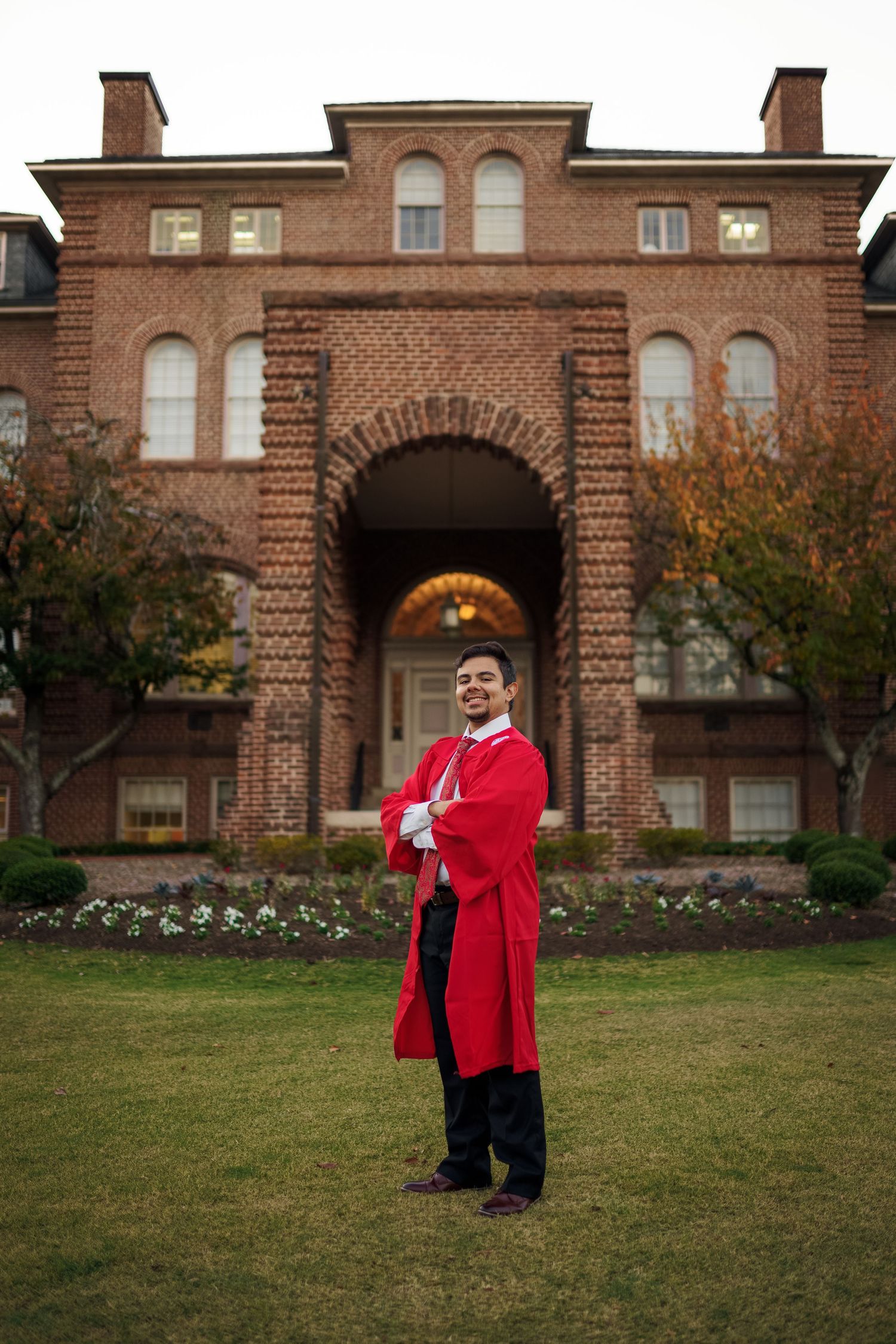 Graduate poses in front of historic brick university building with arched entrance wearing red regalia.