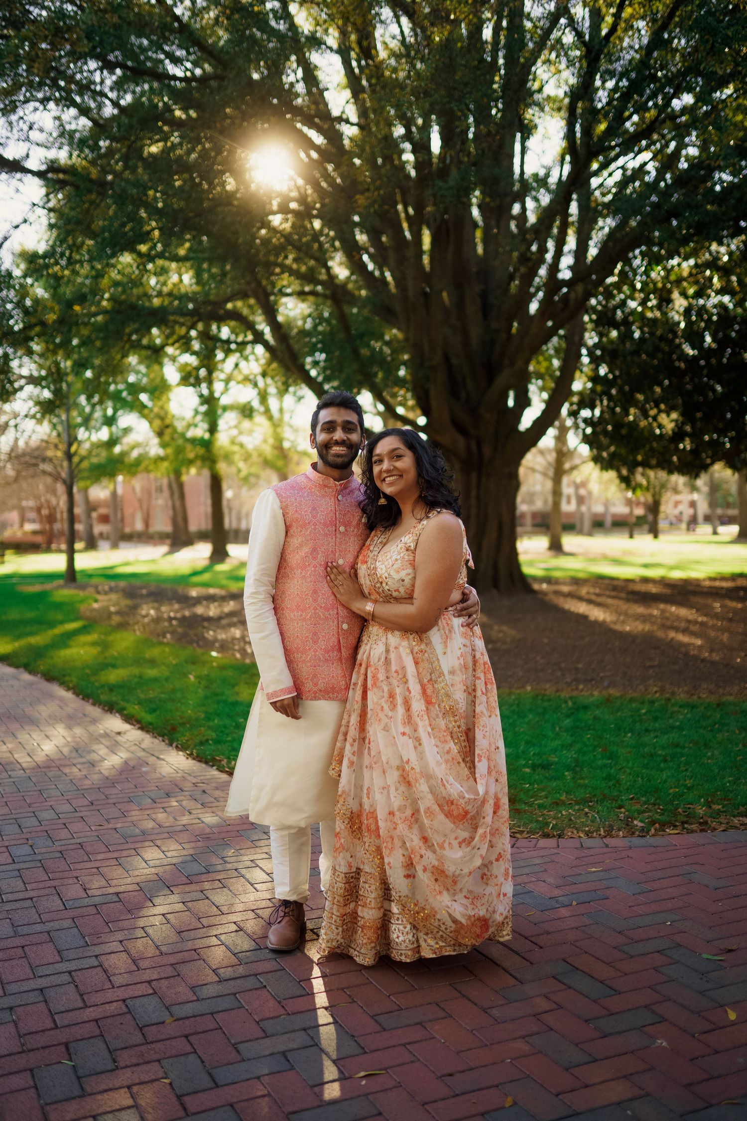 Couple in elegant peach and cream formal attire pose on brick pathway under large tree at sunset.
