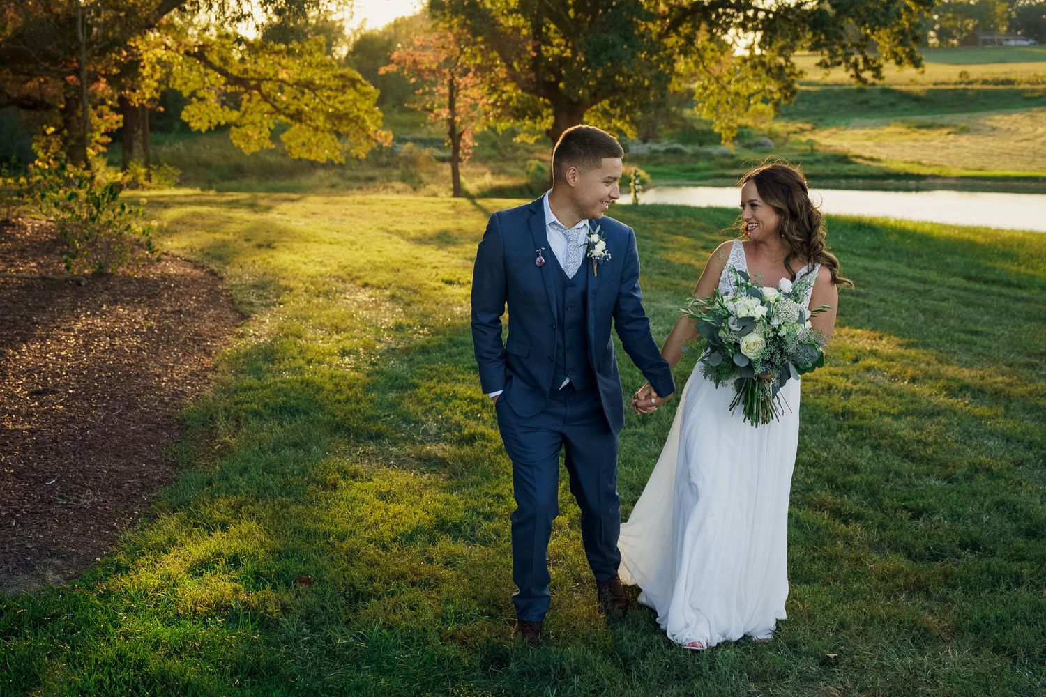 A couple in wedding attire walks together on grass with golden sunlight filtering through trees.