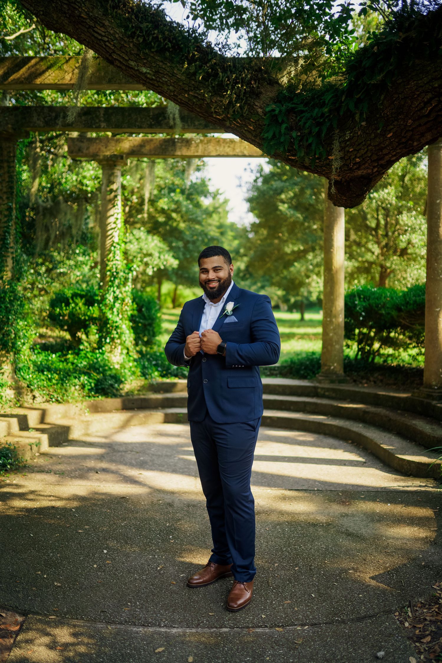 Groom in navy suit poses confidently under garden pergola with natural greenery.
