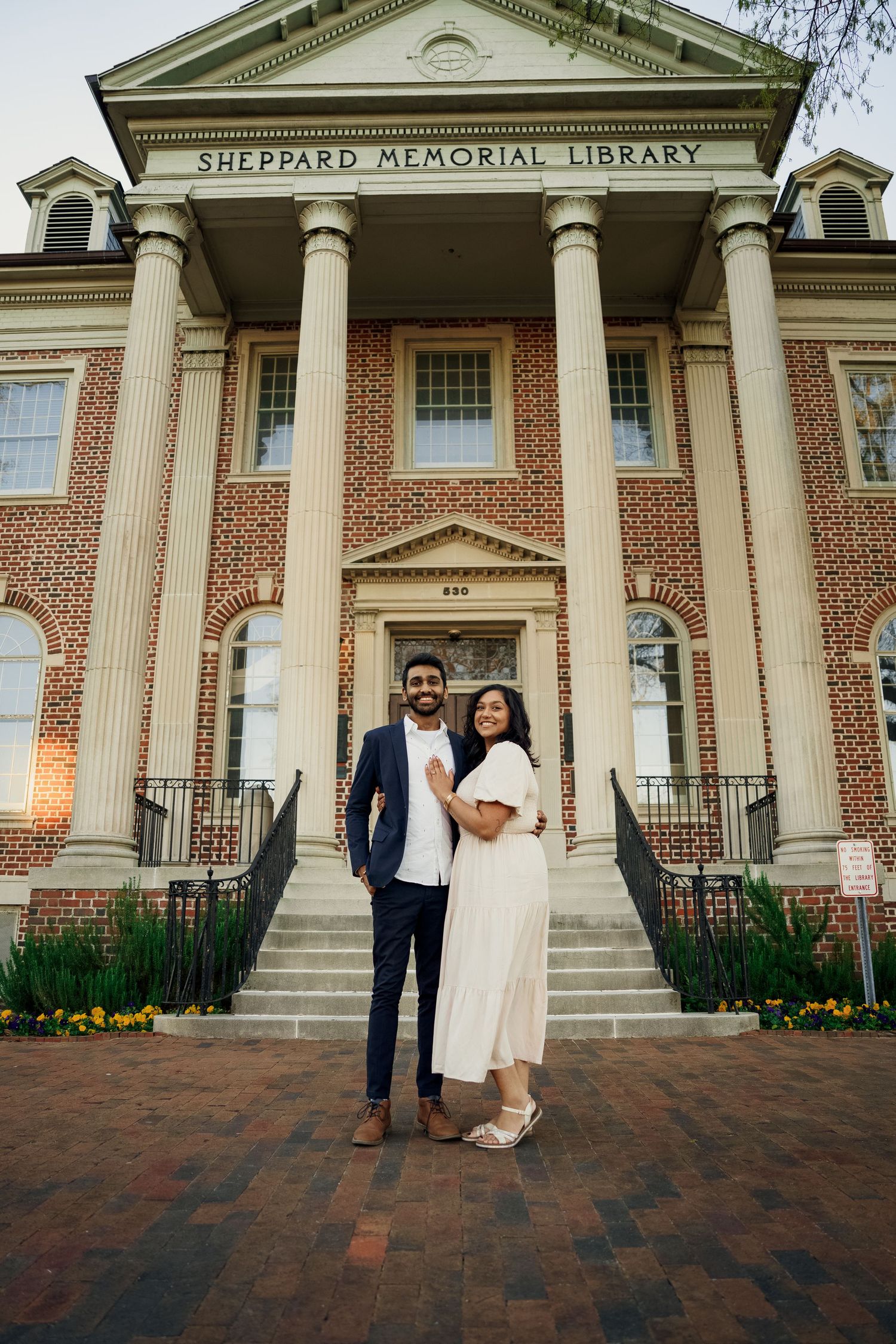 A couple poses in front of Sheppard Memorial Library's classic architecture.