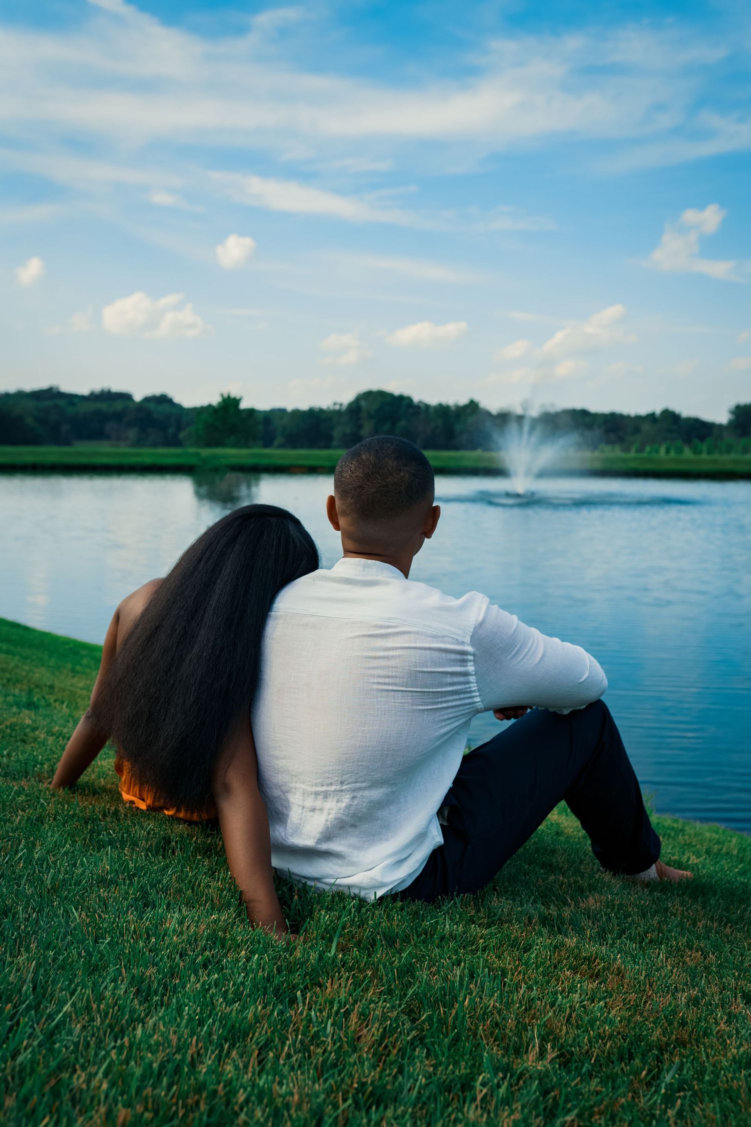 A couple sits by a lake watching a fountain spray water on a sunny afternoon.