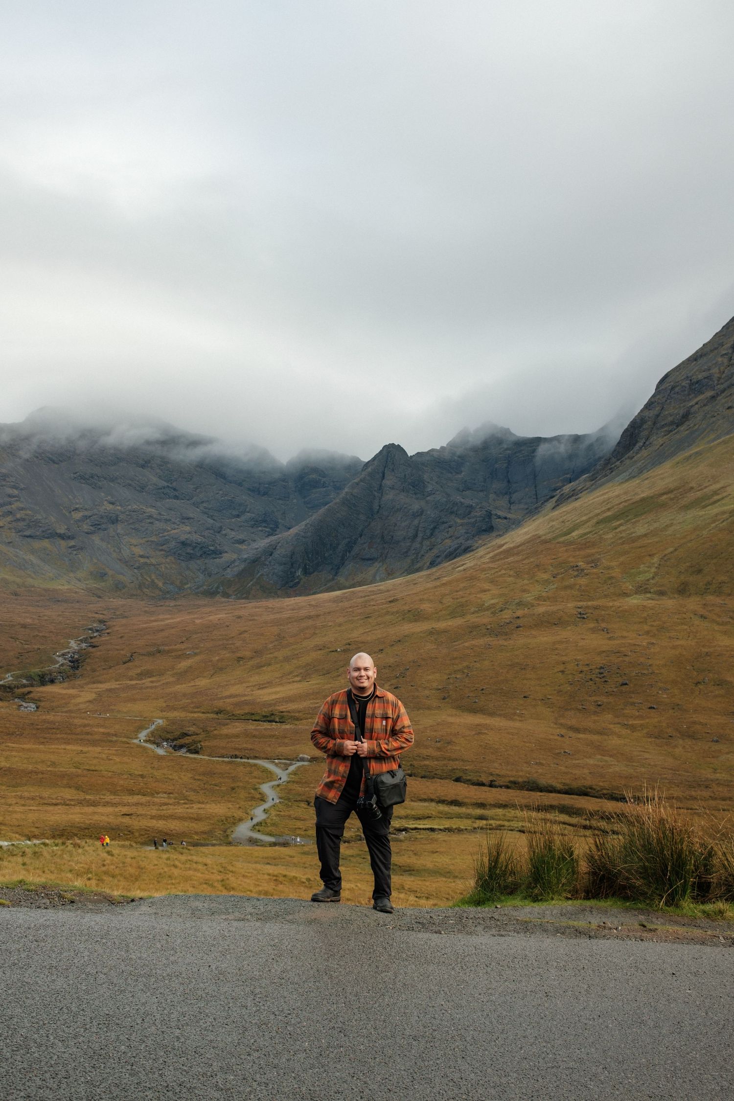 A hiker stands on a mountain road overlooking a misty Scottish valley with peaks in the distance.