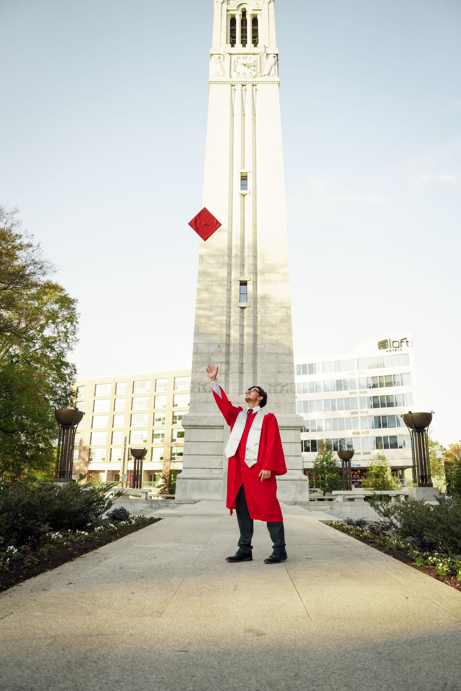Graduate in red regalia tosses graduation cap in front of tall white university bell tower.