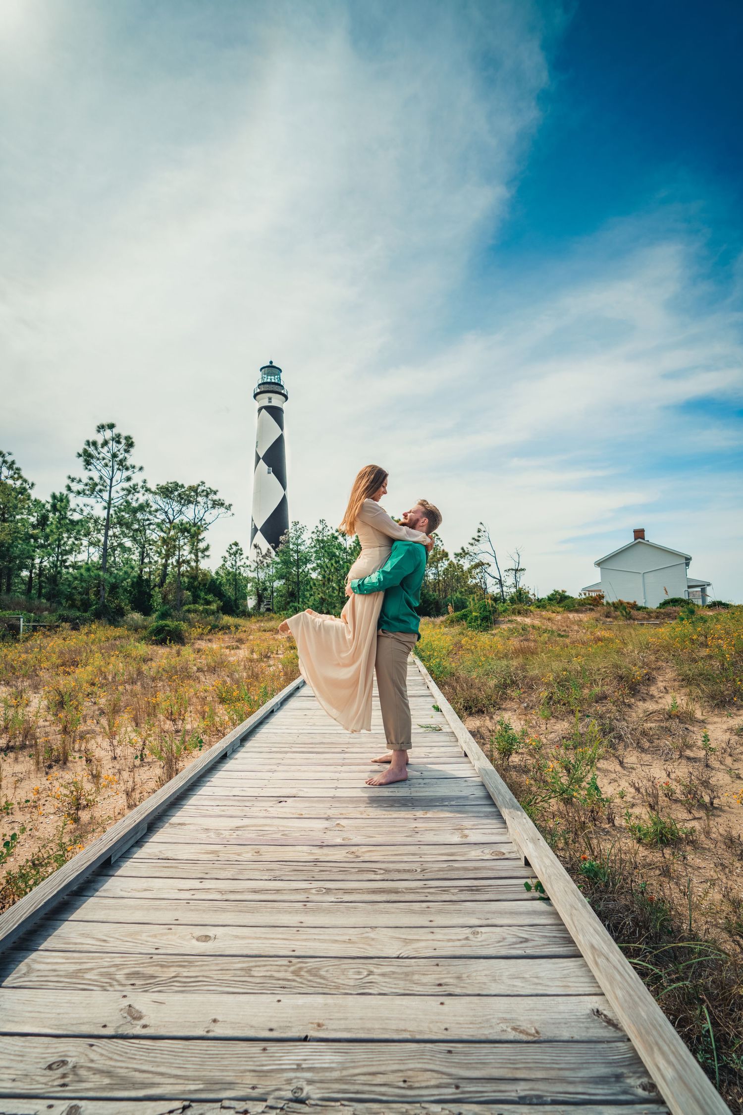 A couple embraces on a wooden boardwalk with the Cape Hatteras lighthouse rising in the distance.