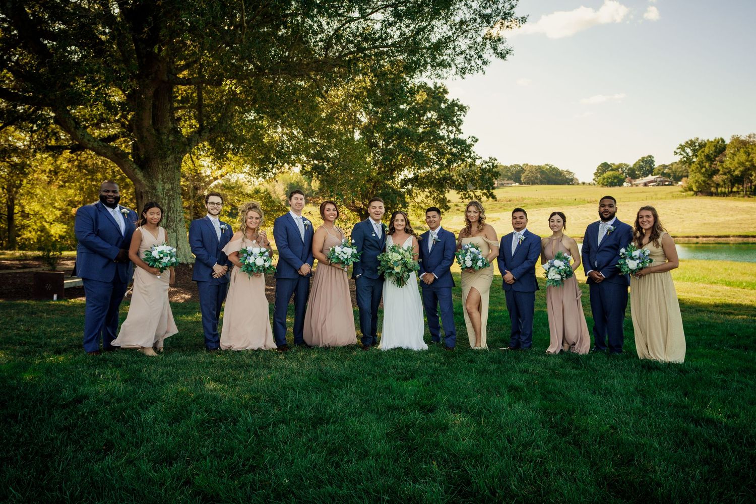 Wedding party lineup in coordinated navy suits and neutral dresses against golden sunset landscape.
