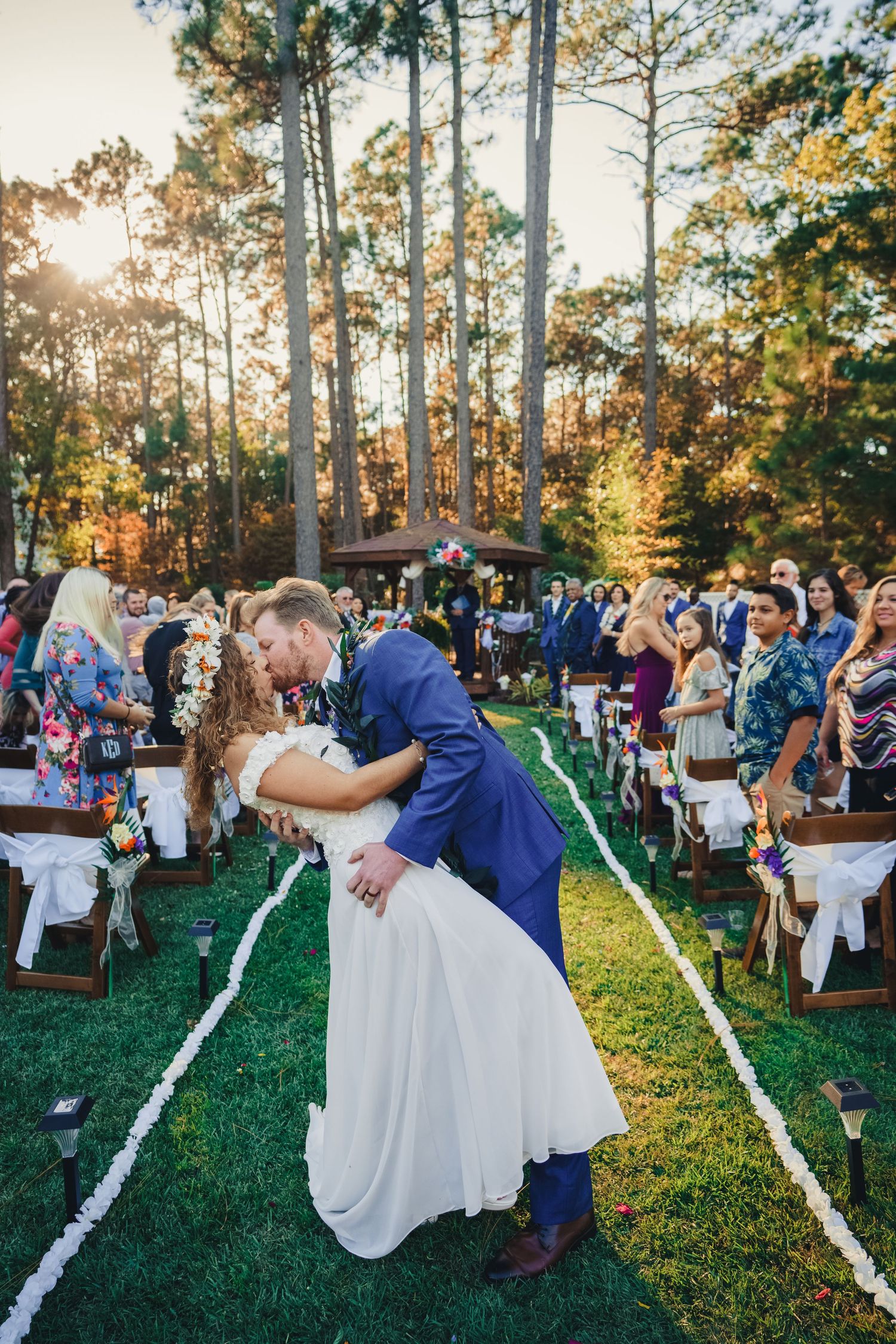 Romantic dip kiss between newlyweds at outdoor forest ceremony with guests looking on.