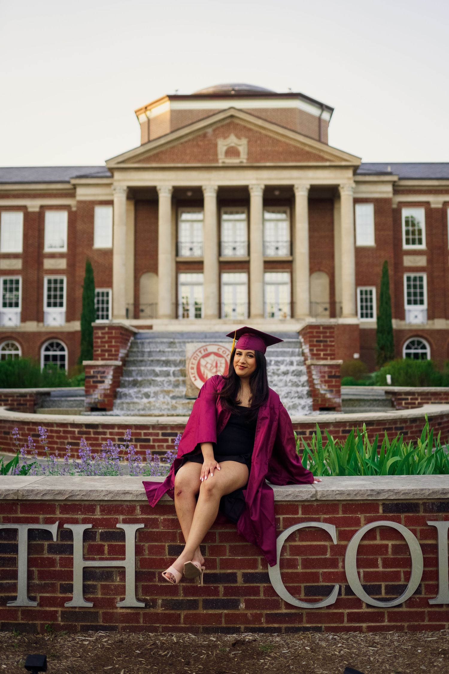 Graduate in maroon cap and gown sits on brick wall at Meredith College campus.