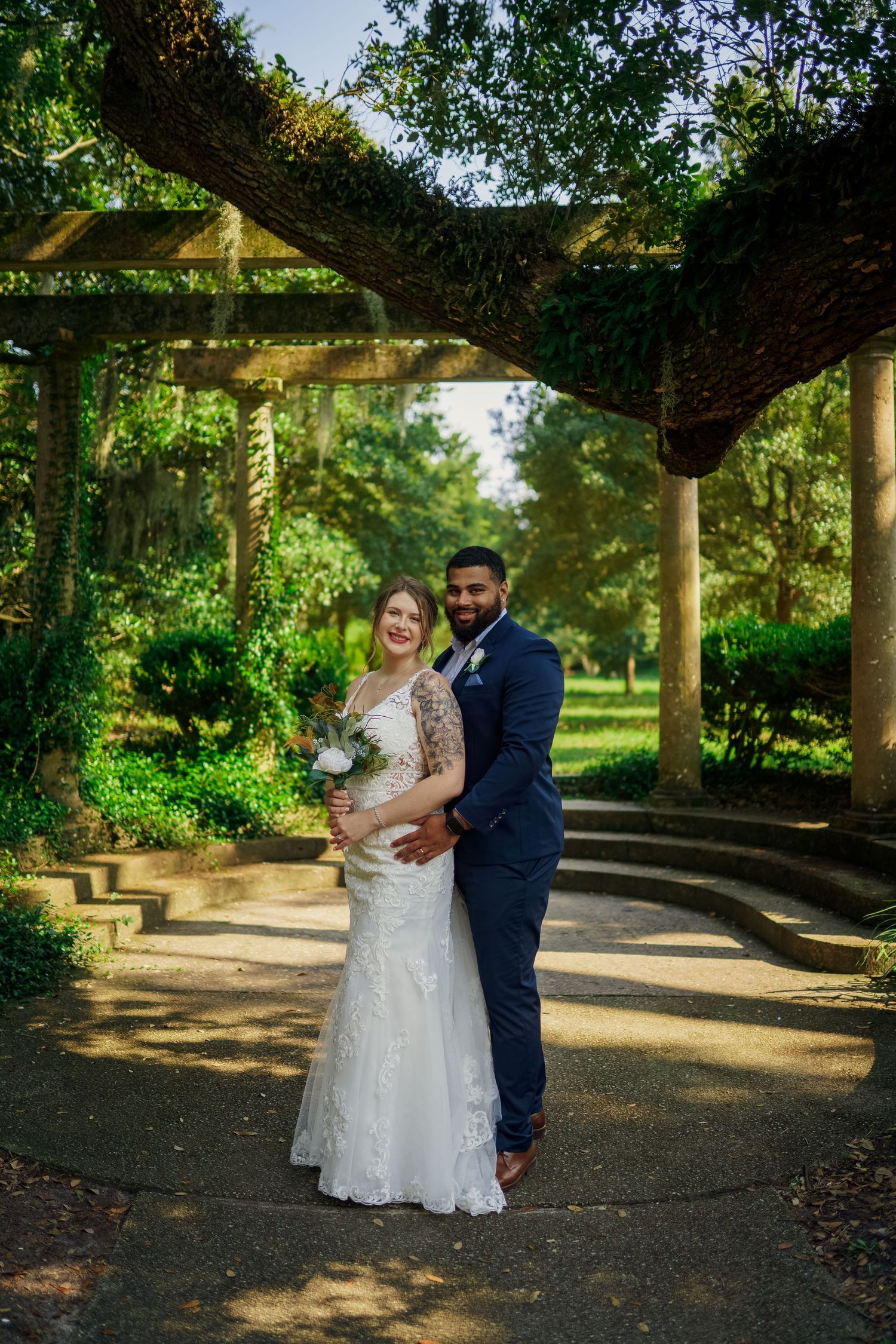 A romantic wedding photo captured under a garden pergola with natural sunlight filtering through the foliage.