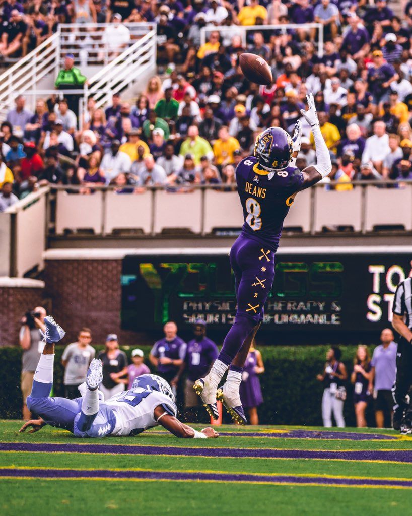 A ECU football player leaps high to catch a pass during a game with a crowded stadium behind over and ECU football player