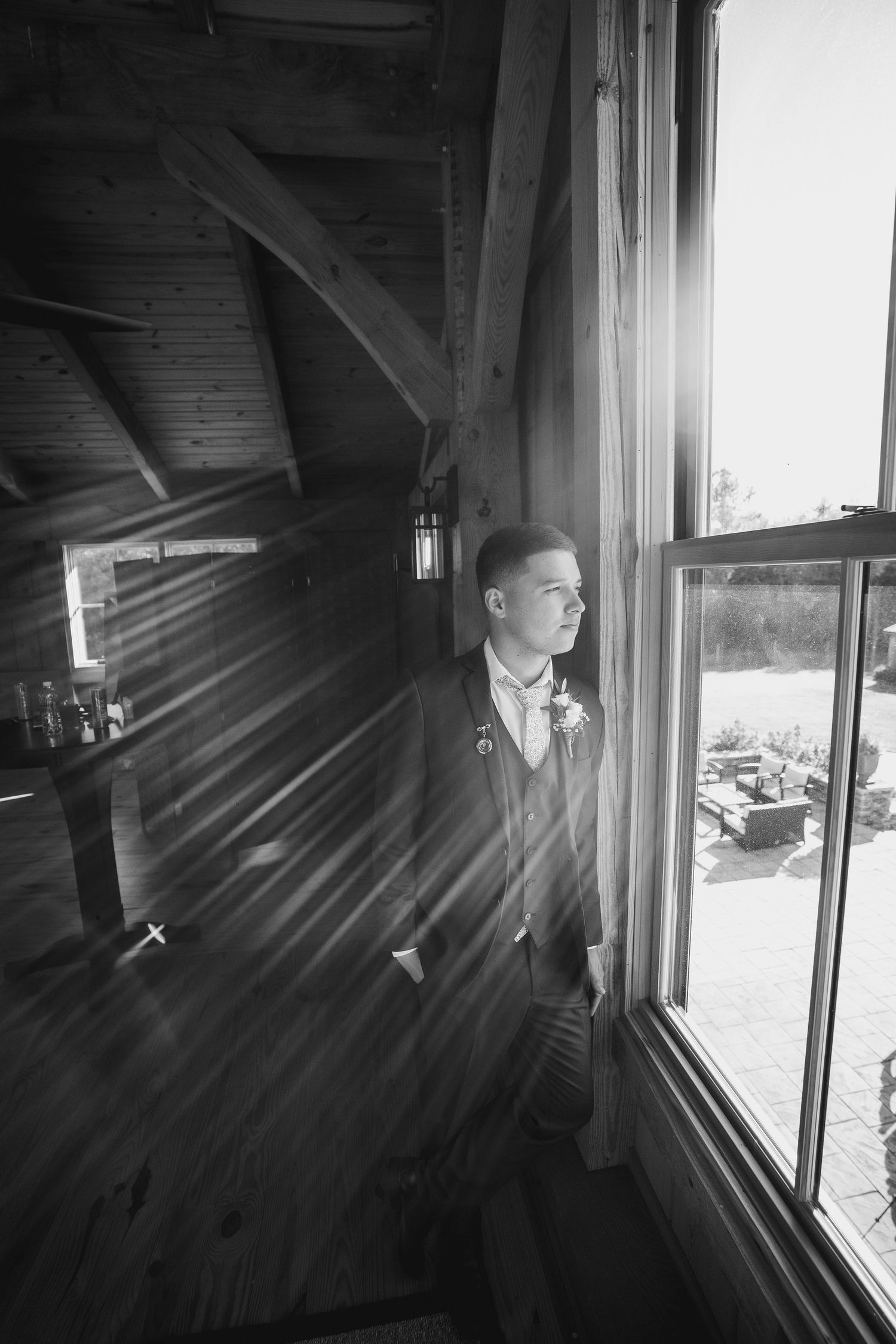 A black and white photo shows a groom in a suit gazing thoughtfully out of a rustic cabin window.
