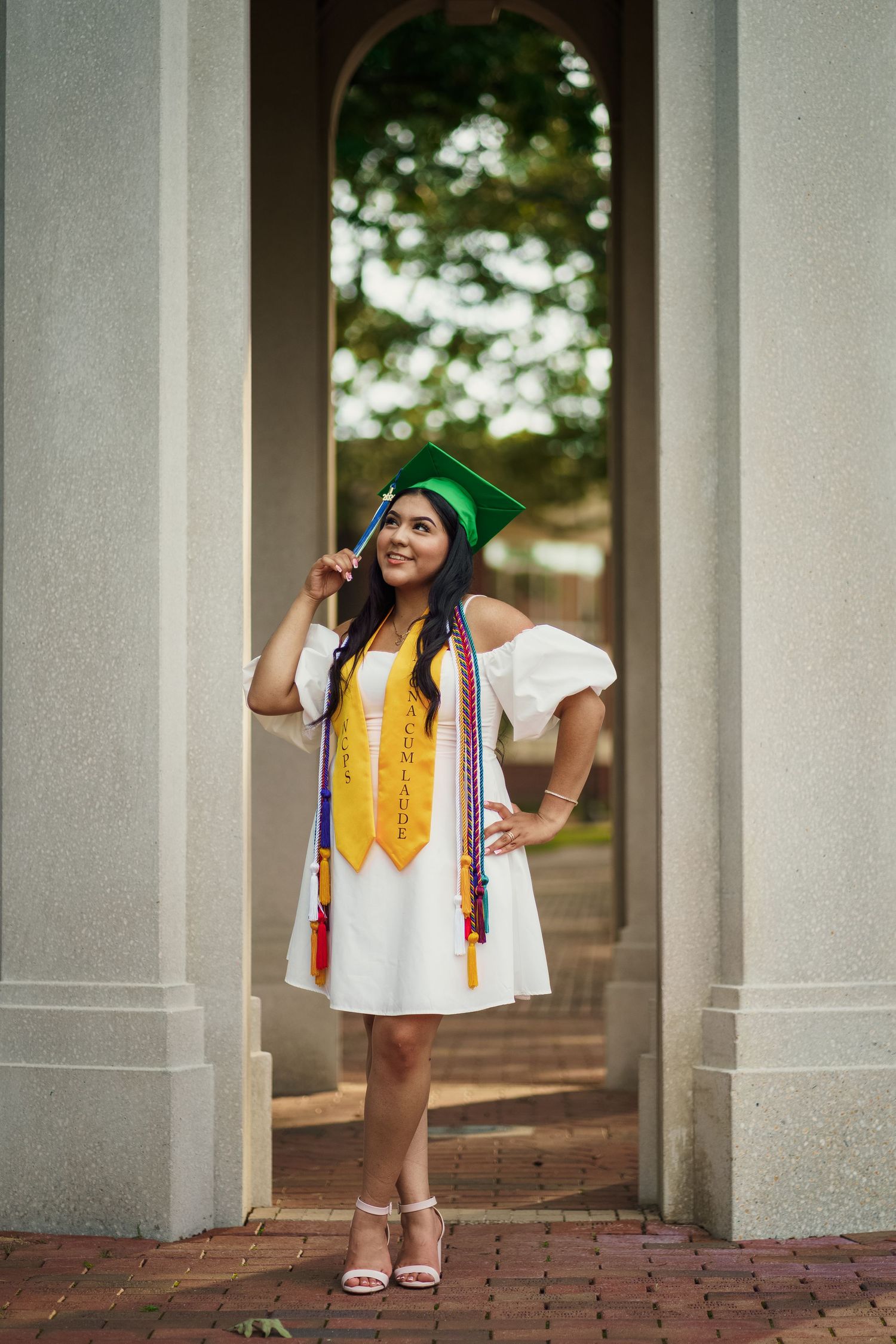 Graduate in white dress and green cap poses under archway with graduation stole and cords.