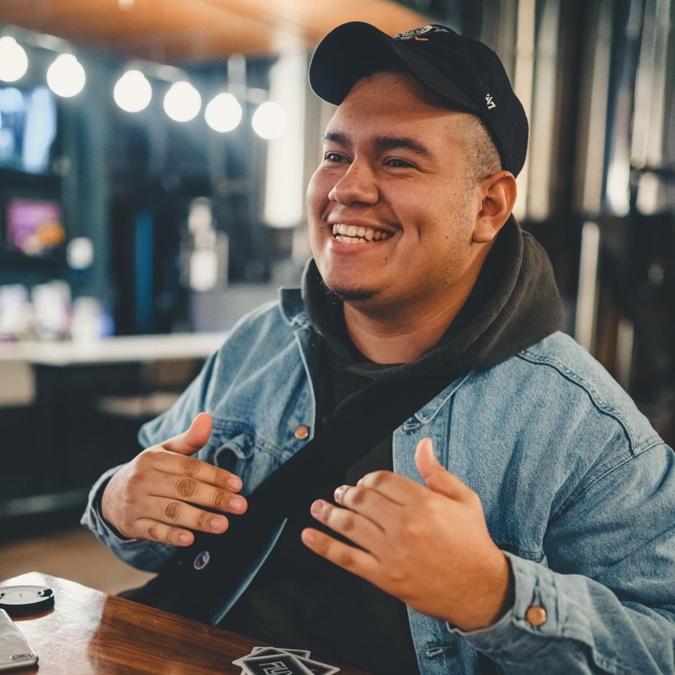 A person wearing a baseball cap and denim jacket smiles while sitting at a table with bokeh lights behind.