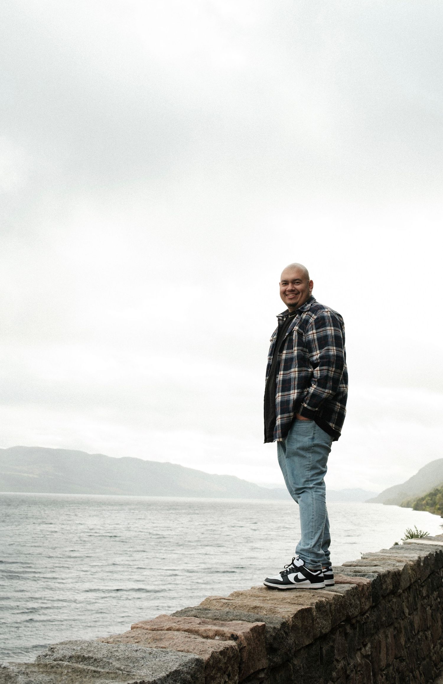 A person in plaid stands on a stone wall overlooking a misty coastal landscape at Loch Ness