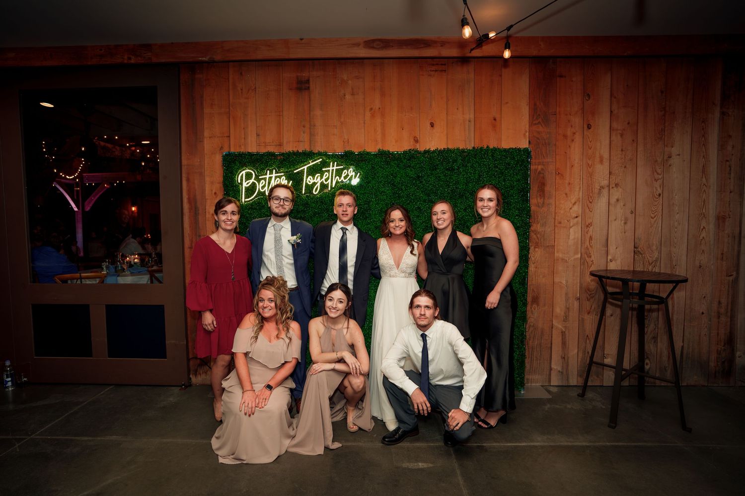 Wedding party poses against wooden wall with neon sign in rustic venue.
