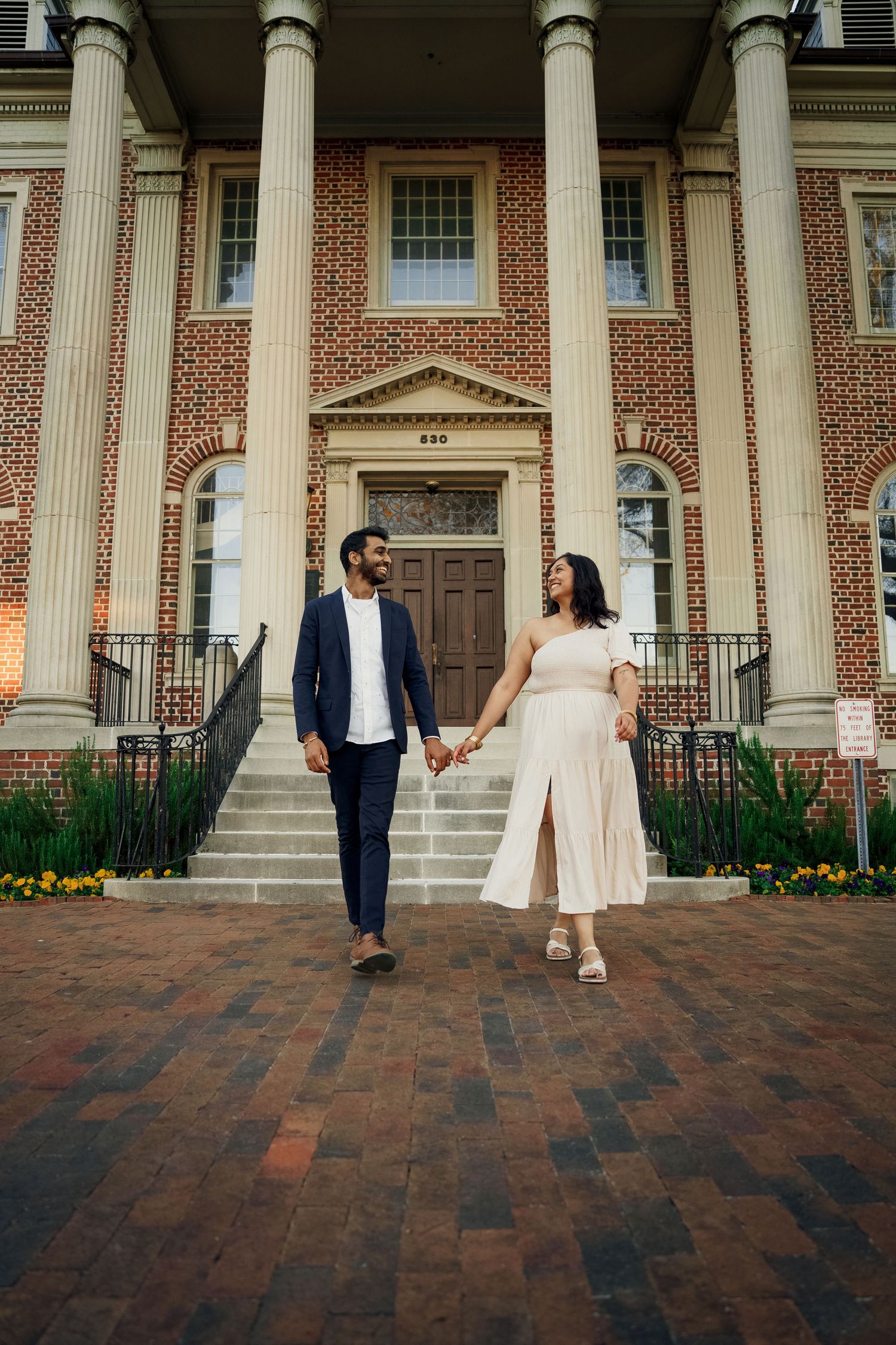 A couple walks hand in hand in front of a grand colonial-style building with columns.
