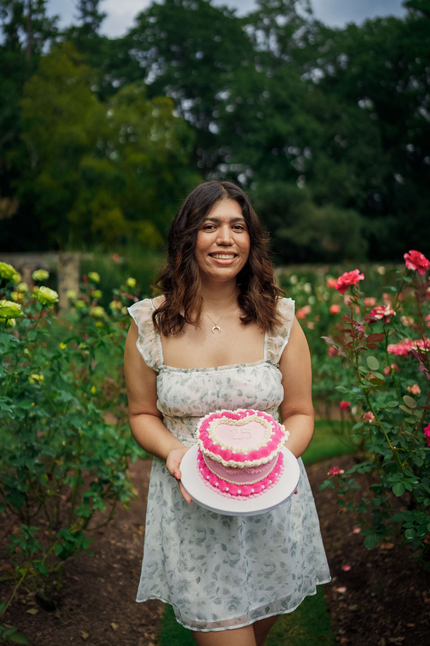 Person in floral dress holds pink birthday cake in blooming rose garden.