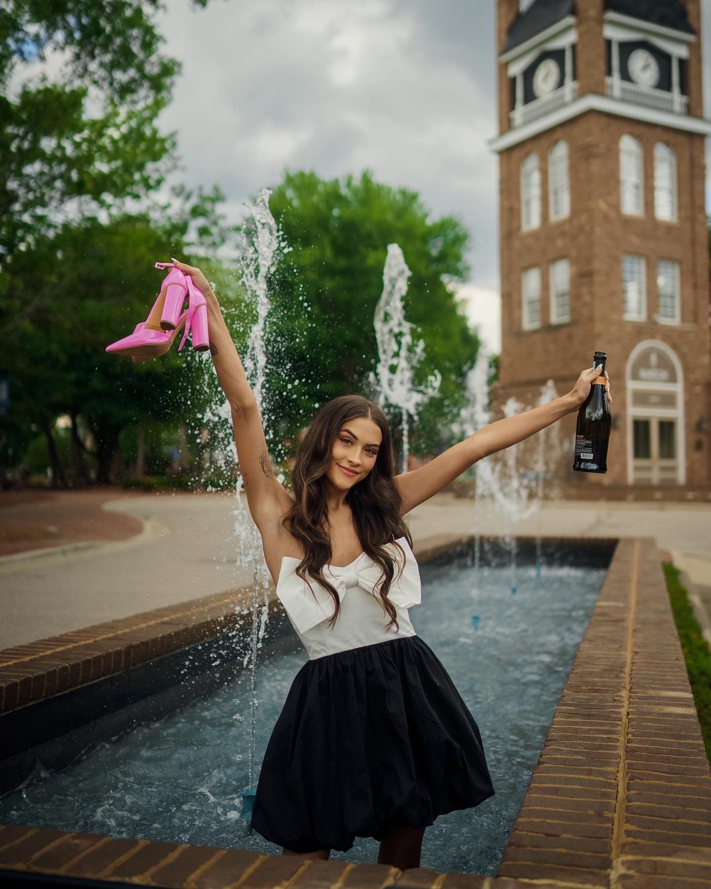 Barton graduate celebrates joyfully by a fountain while holding up shoes and a bottle near a brick clock tower building.