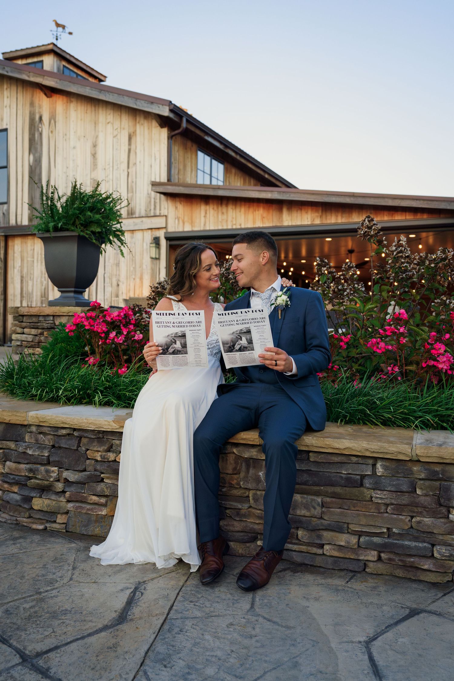 Couple in wedding attire holding newspapers while sitting on stone wall by rustic barn.