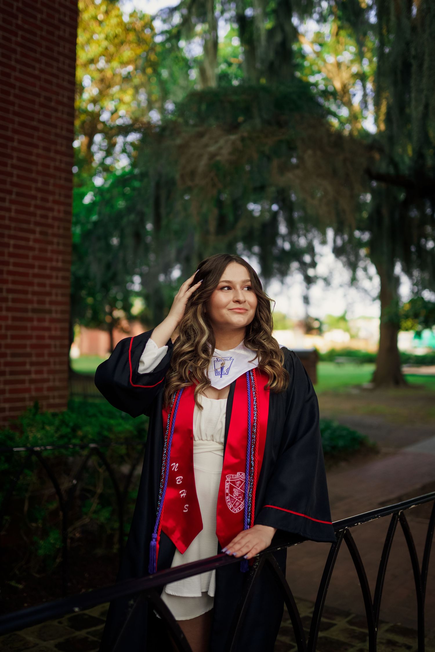 Graduate in academic regalia with red stole poses near brick building.