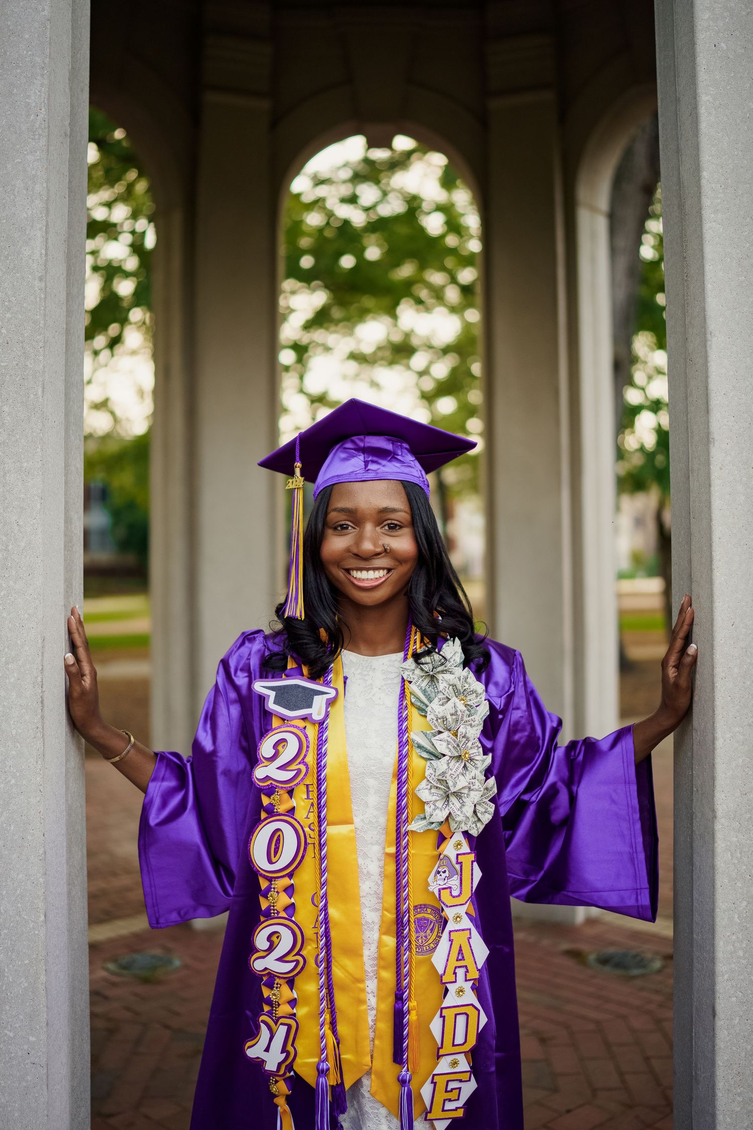 Graduate in purple cap and gown with decorative sash and honor cords stands smiling in the ECU Cupola while she poses for grad photos.