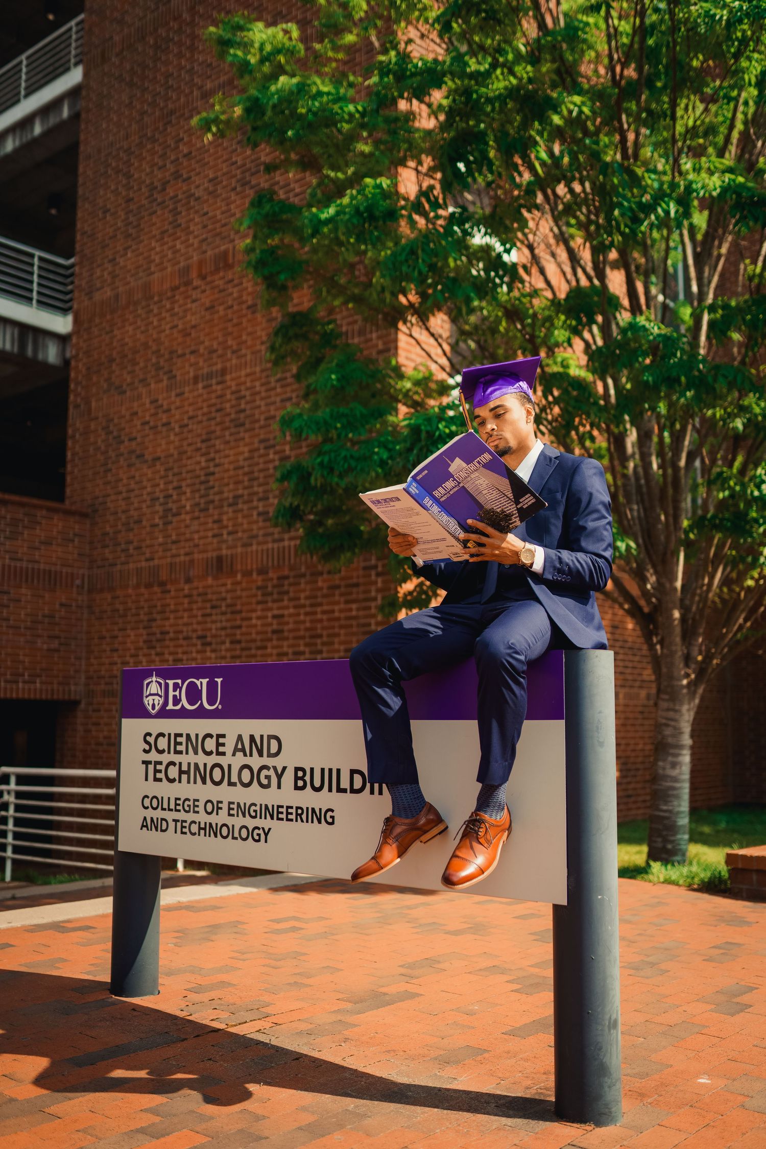 Graduate in cap and gown reading a book while sitting on the Sci tech sign outside ECU Science and Technology Building.
