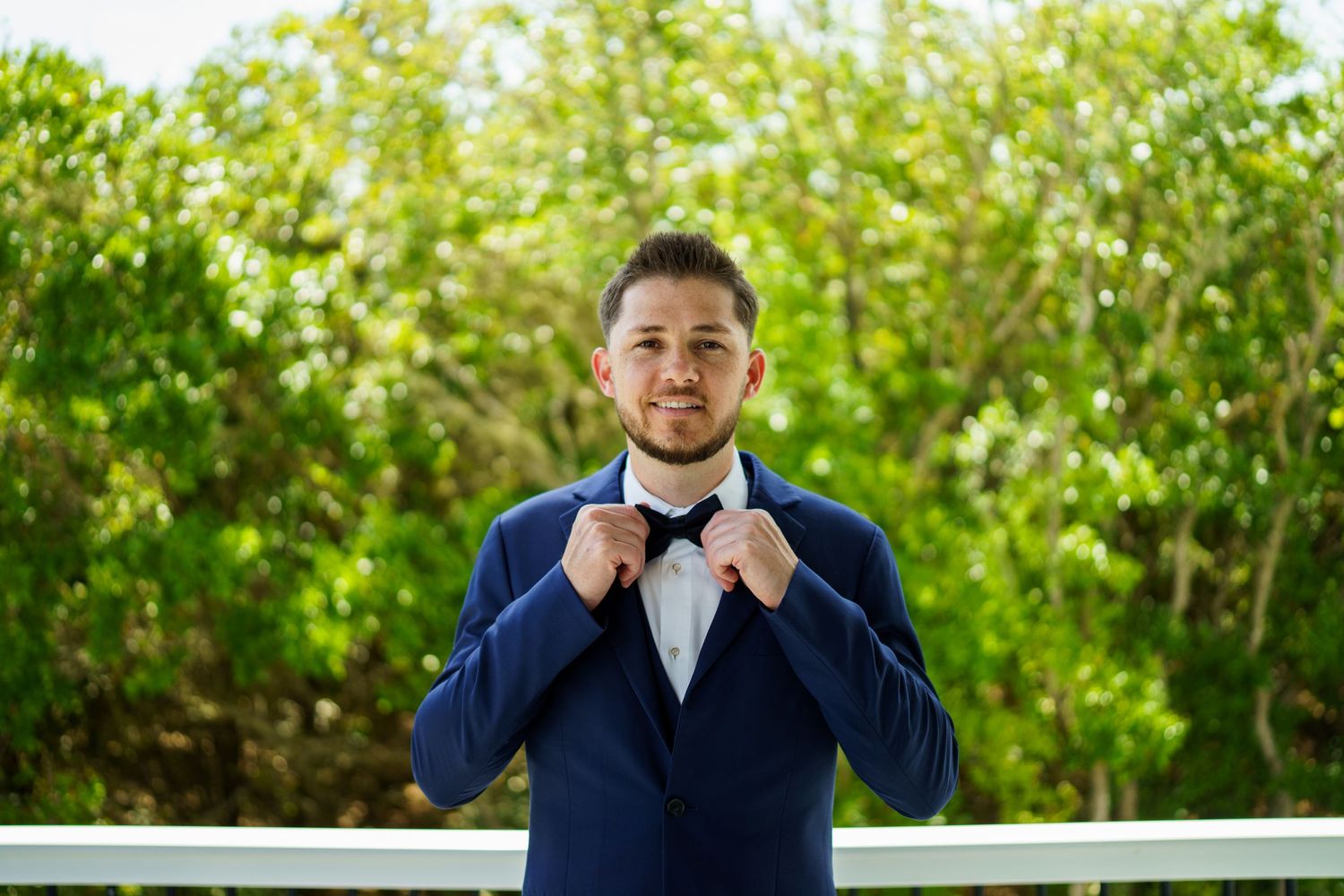 A person in a navy blue suit adjusts their bow tie while standing outdoors against a green leafy background.wedding photographer Joan Avilez