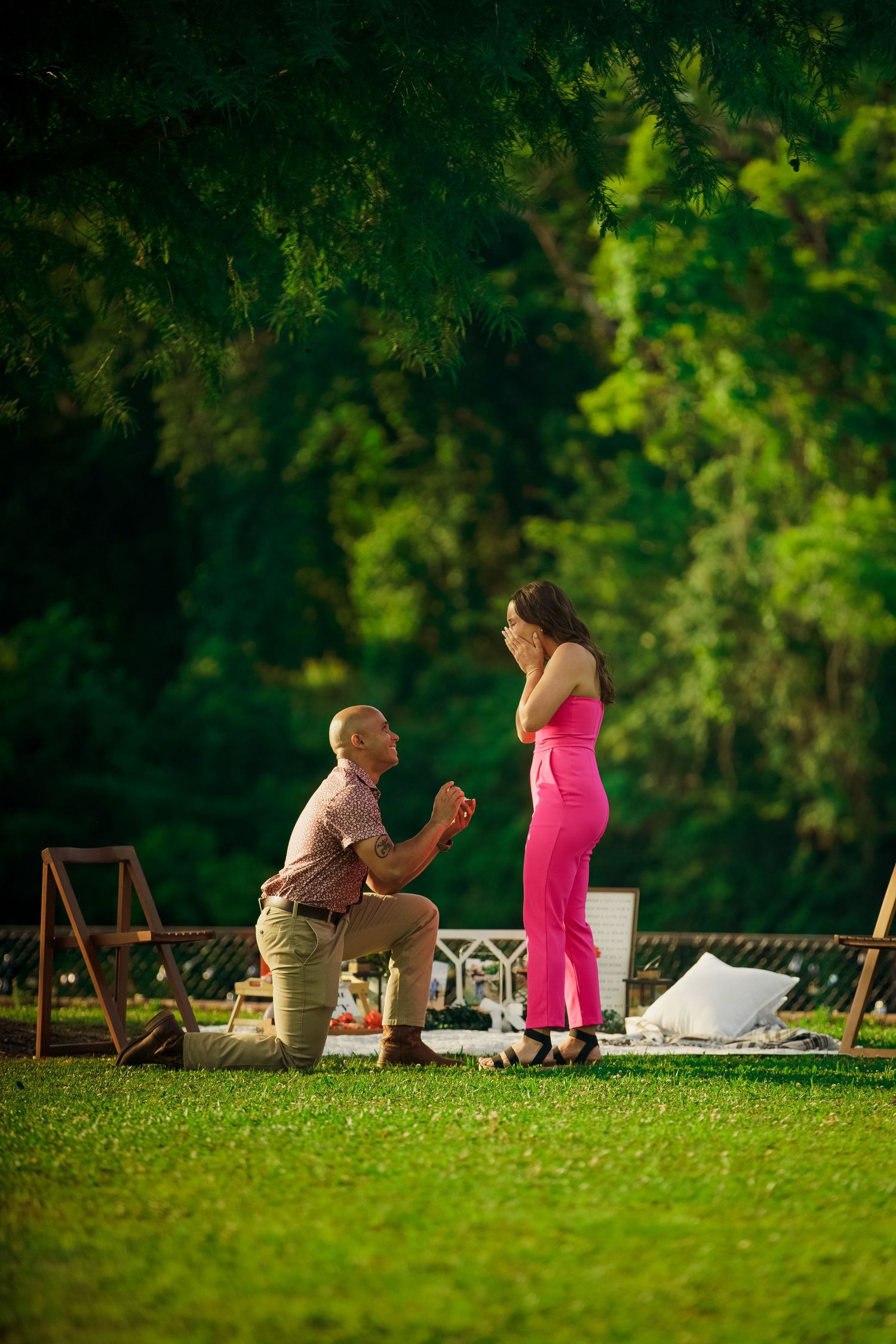 A romantic outdoor proposal scene set up on a lawn with wooden chairs and greenery.
