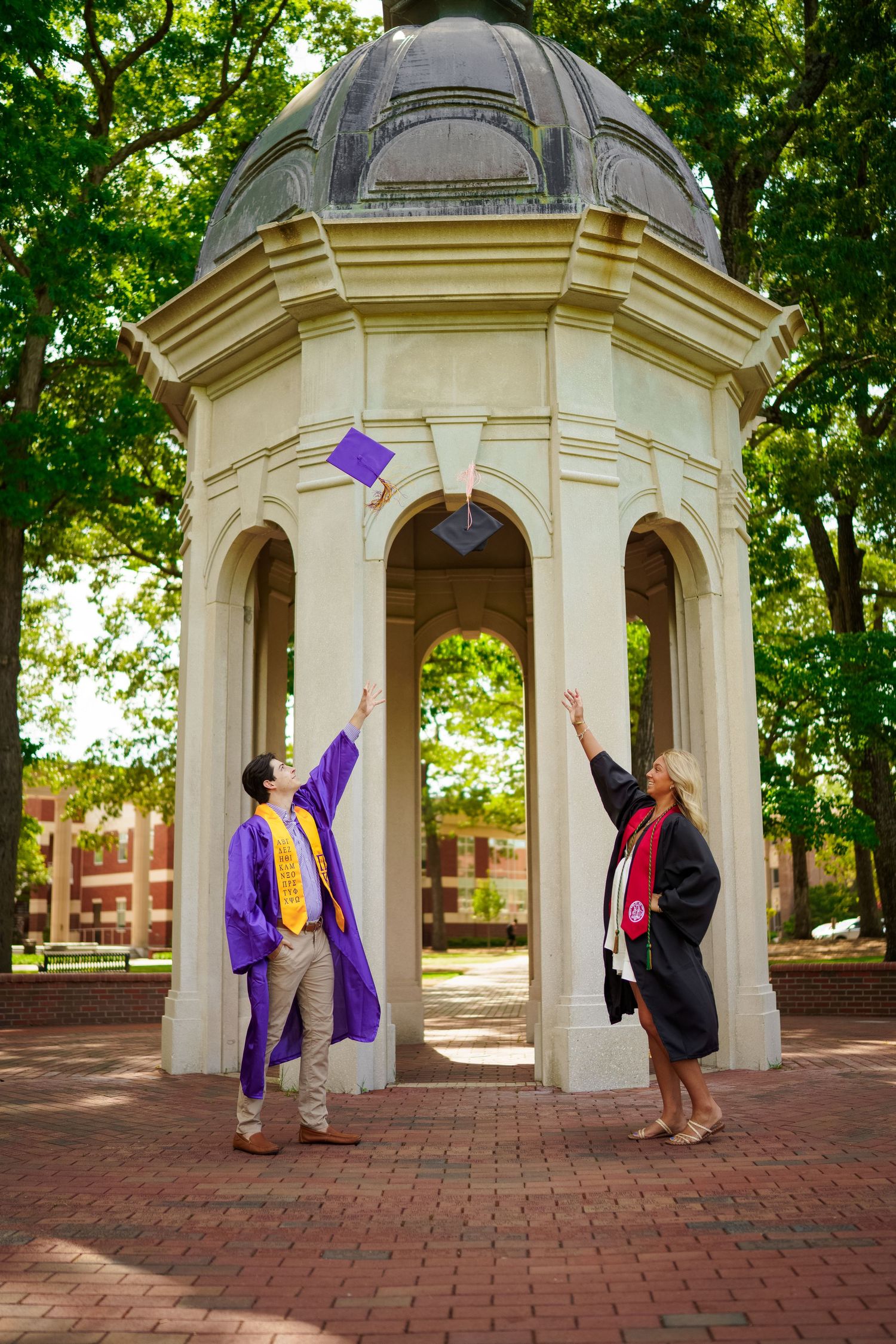 graduates in caps and gowns toss their graduation caps into the air under a ECU Cupola on a brick patio. Joan Avilez ECU grad photographer