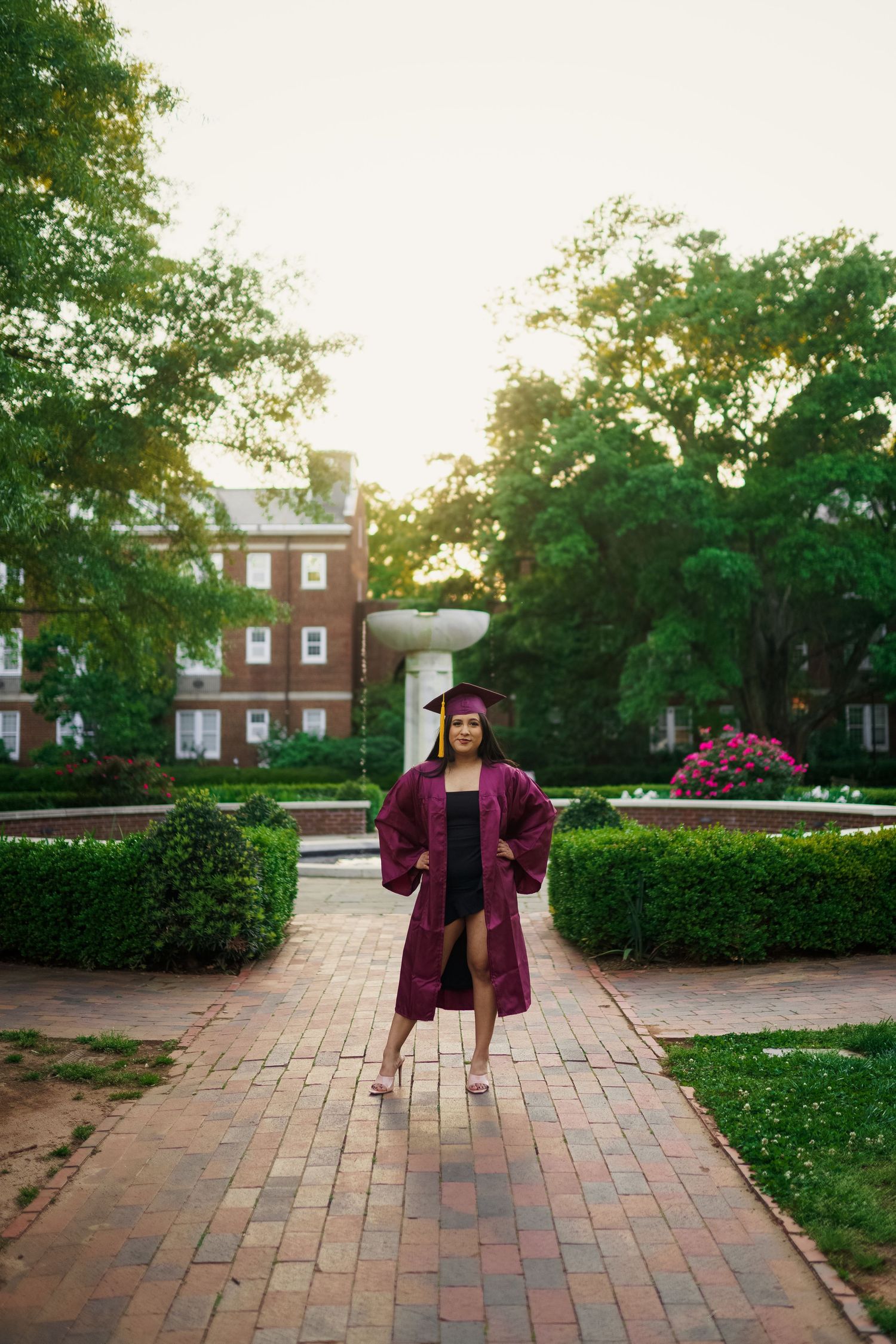 Graduate in maroon academic regalia stands on brick pathway with campus building in background.