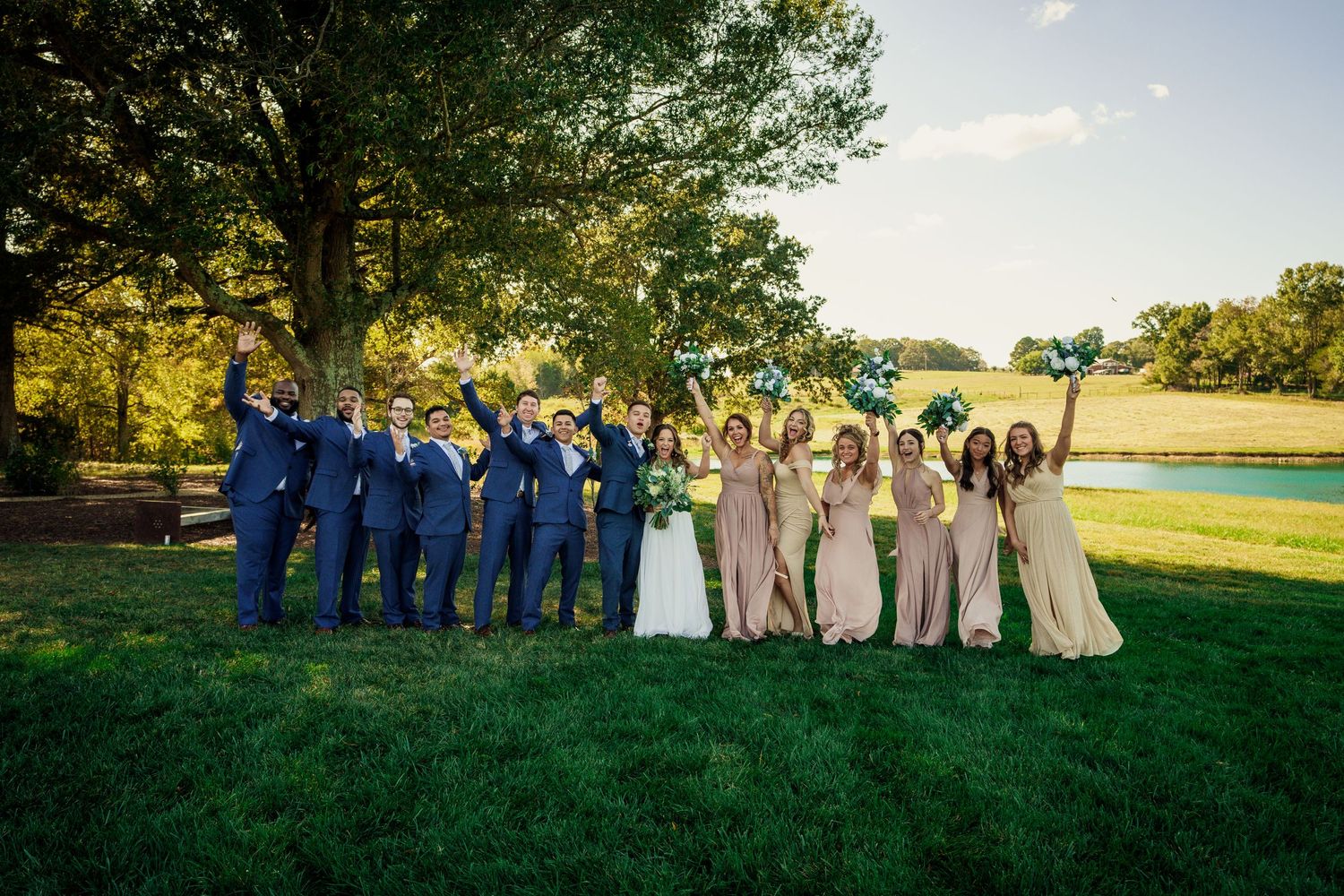 Wedding party in navy and beige poses on grass with trees and setting sun in background.