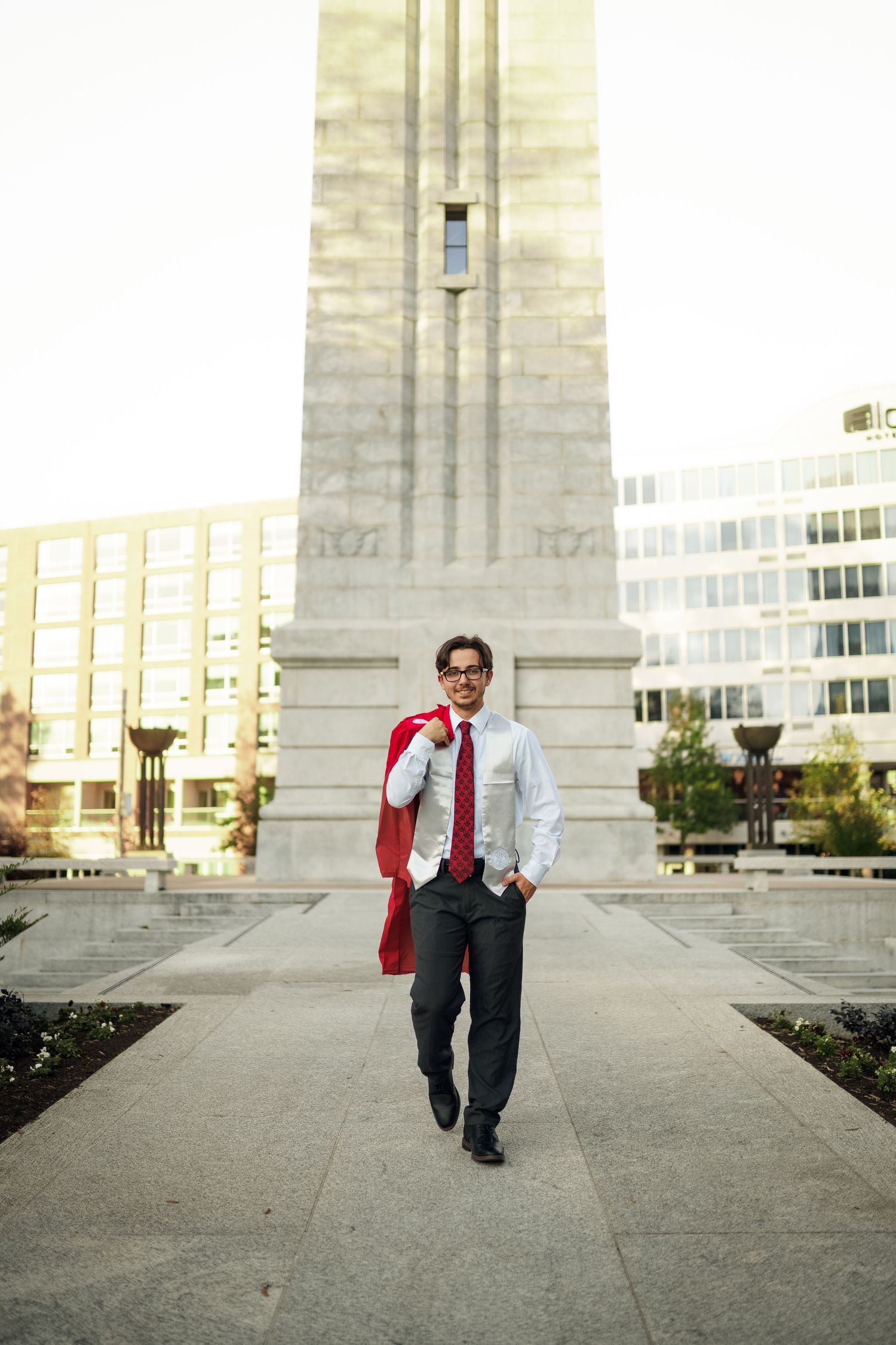 Graduate in red academic regalia walks confidently in front of towering concrete university bell tower.