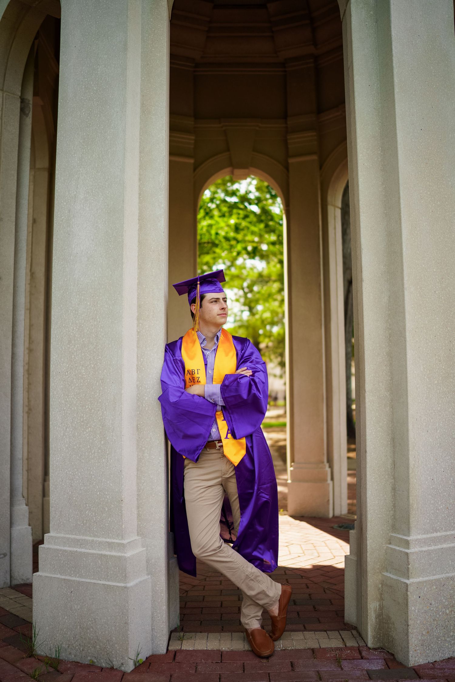 ECU Graduate wearing purple and gold graduation regalia poses casually between classical columns at ECUs cupola with joan avilez photo.