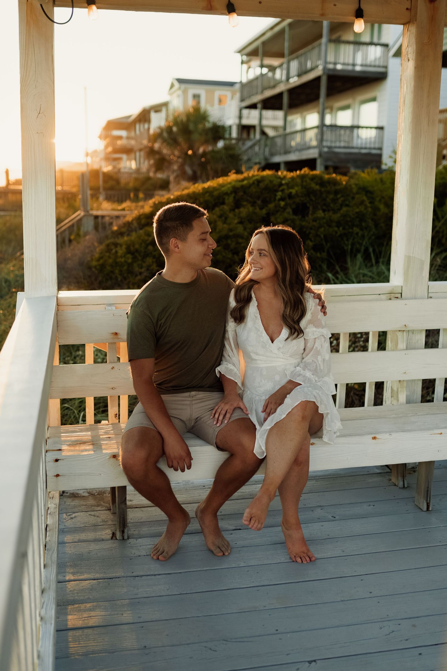 A couple sitting on a white porch railing at the beach during sunset.