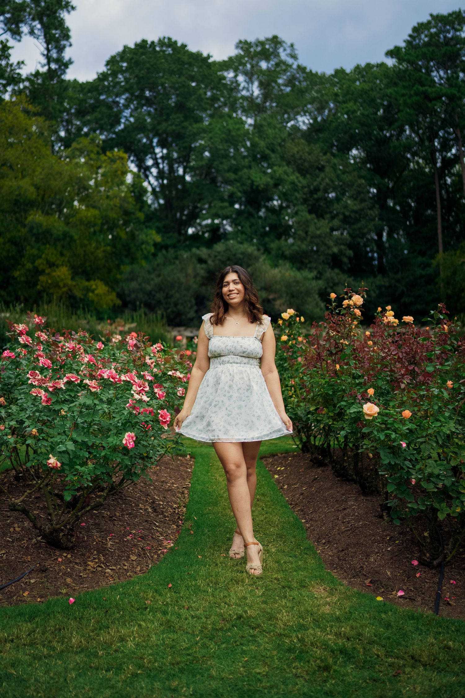 Person in white summer dress poses in beautiful rose garden pathway.